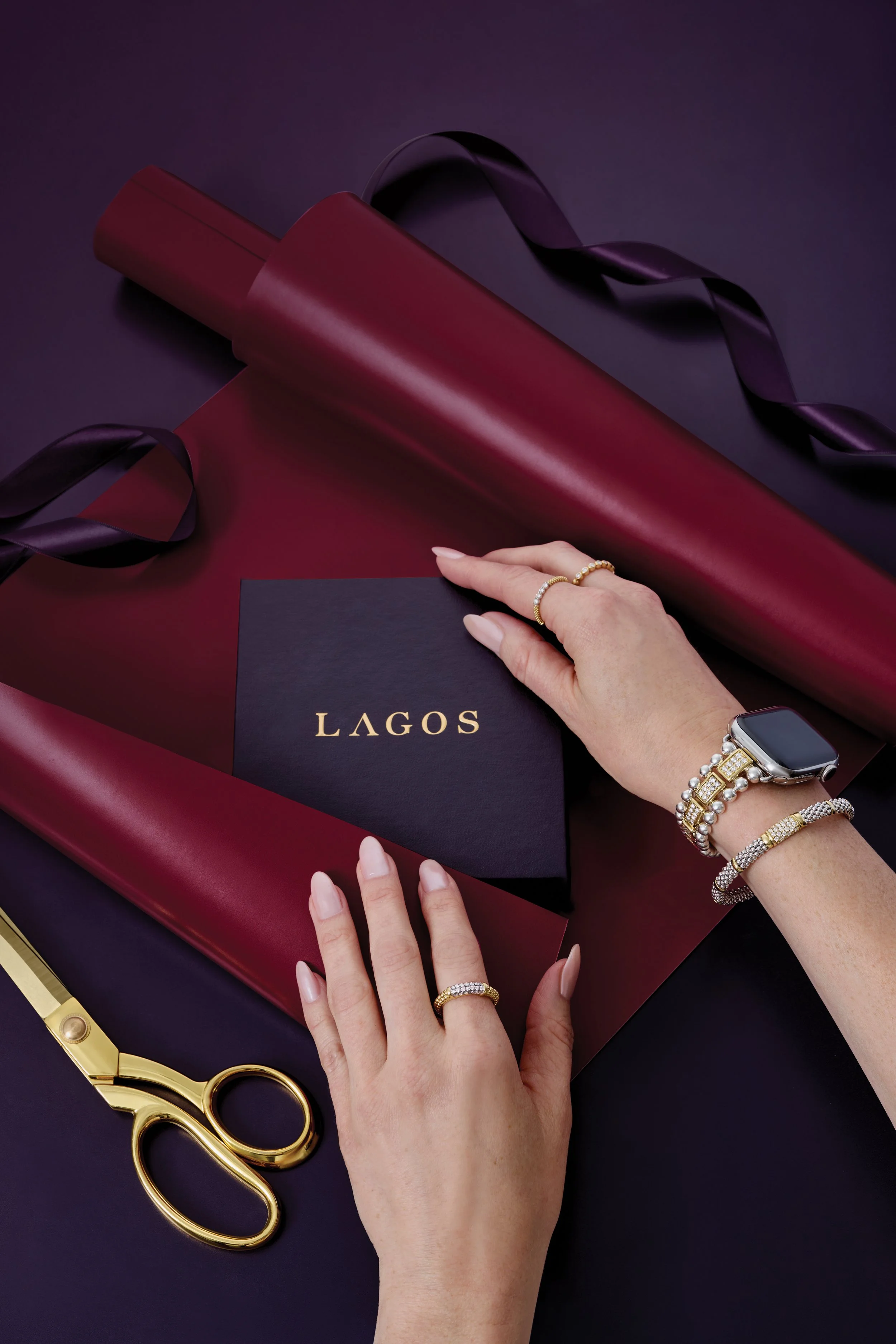 A person's hands arranging black paper with the word 'LAGOS' on it, surrounded by gold scissors, jewelry, and dark red wrapping paper with black ribbon.