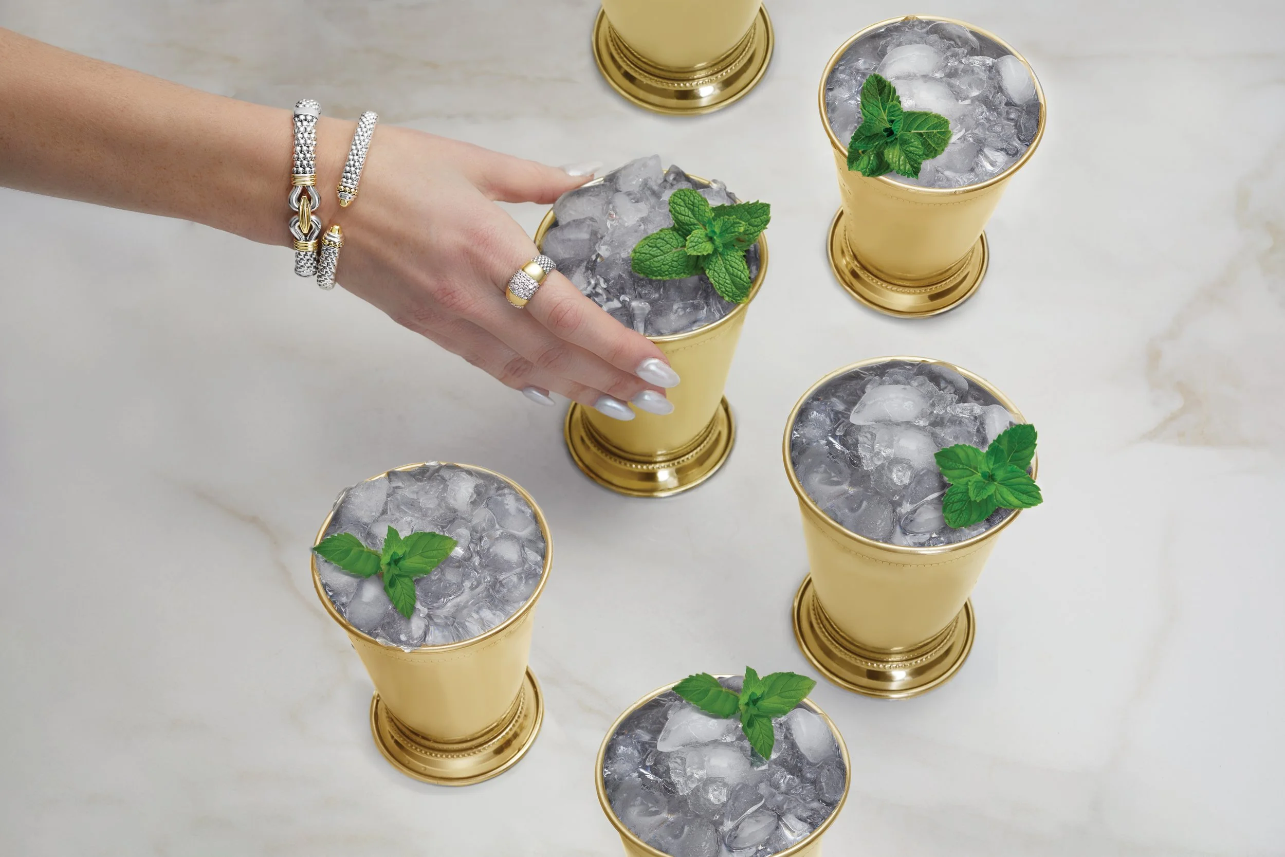 A hand with jewelry arranging five glasses filled with ice and garnished with mint leaves on a marble surface.