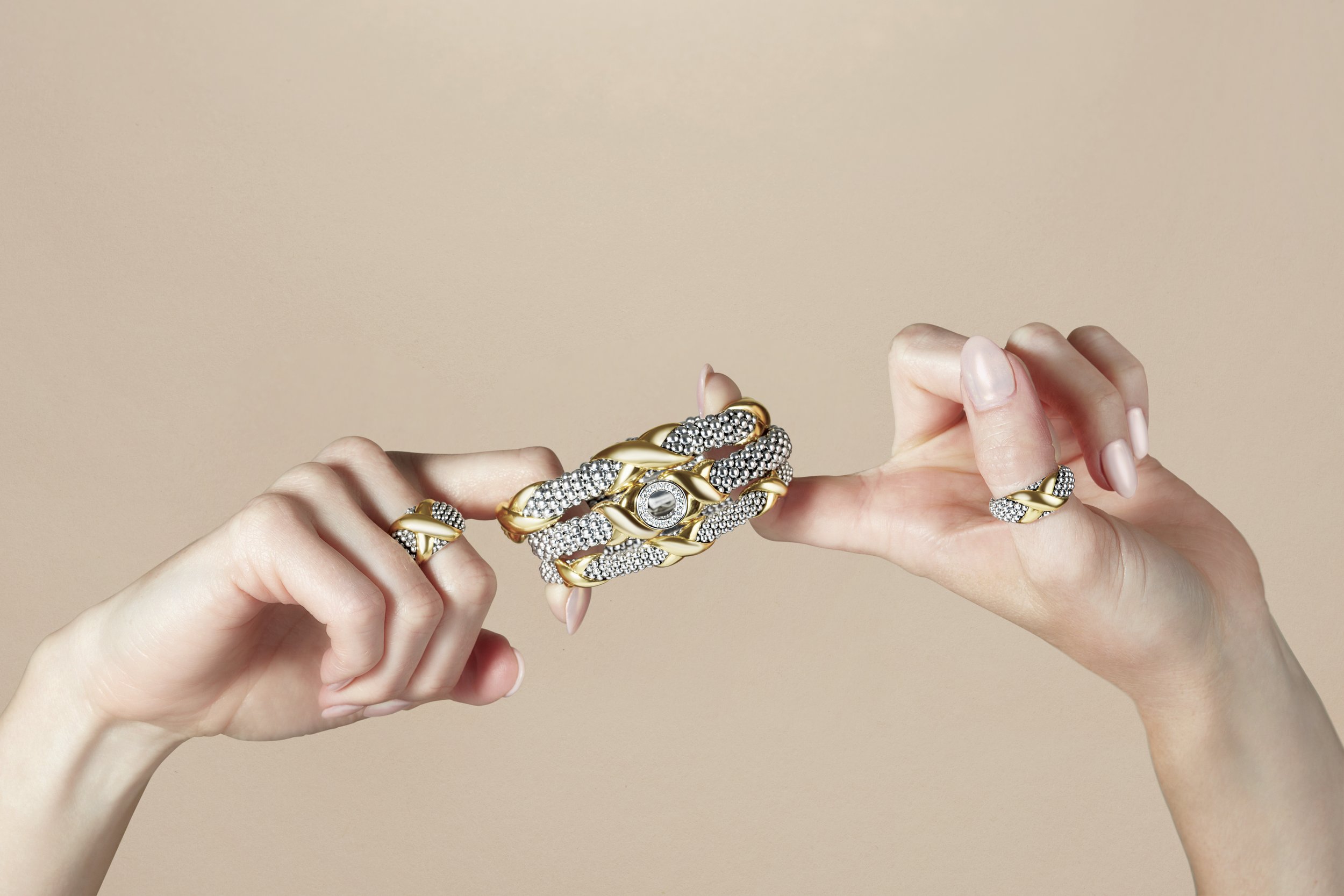 Two hands holding and showcasing a set of gold and silver jewelry, including rings and a bracelet, against a neutral beige background.