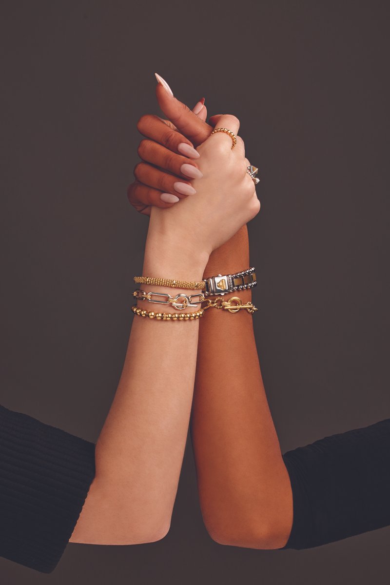 Two hands clasped together, one with darker skin and the other with lighter skin, both wearing multiple gold and silver bracelets and rings, against a dark background.