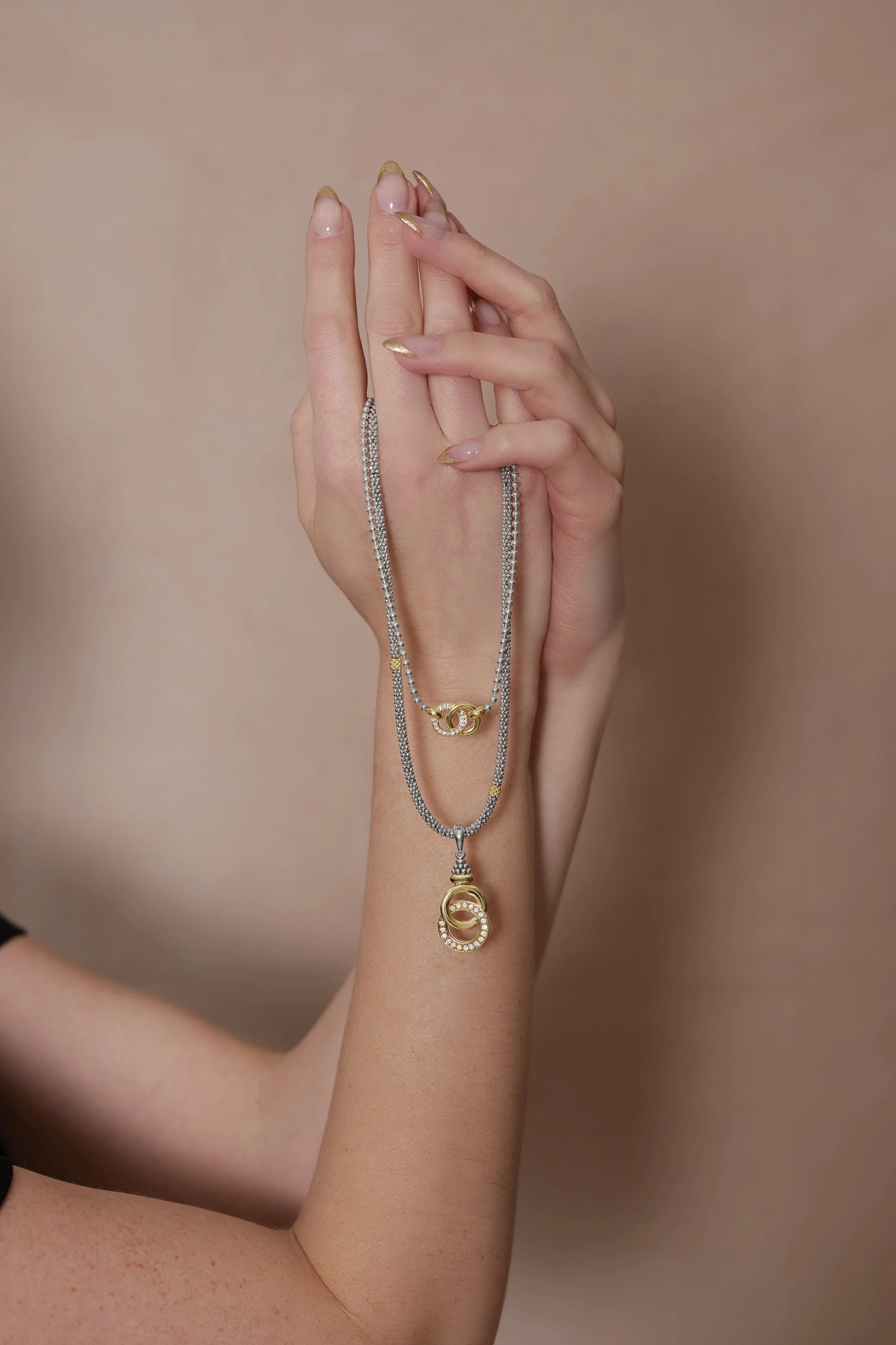 Two hands holding layered silver and gold necklaces with decorative pendants against a beige background.