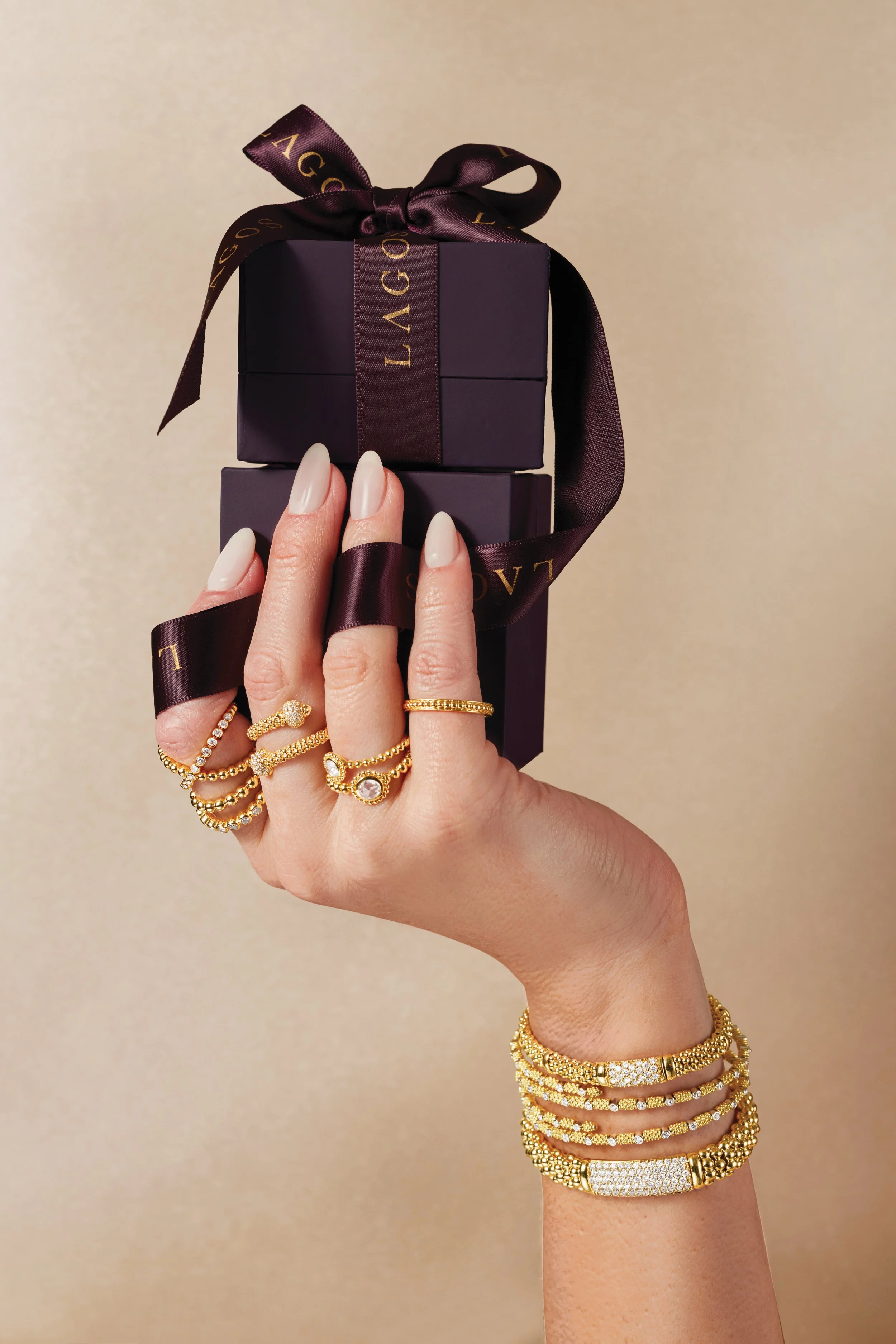 A hand adorned with multiple gold rings and bracelets holds a small purple jewelry box with a ribbon.