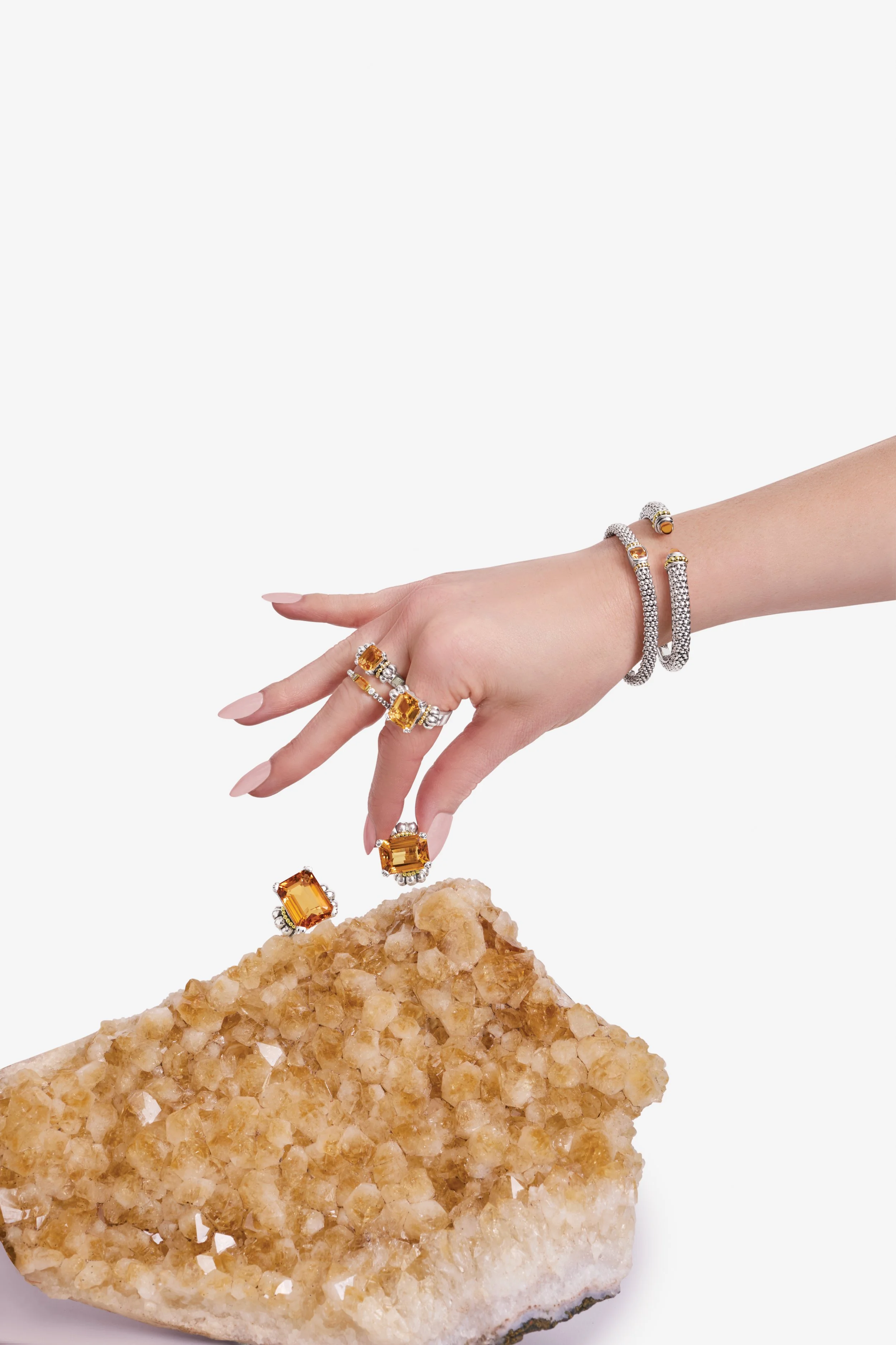 A hand with multiple rings and bracelets is reaching towards a large, yellow gemstone and diamond jewelry pieces resting on a quartz crystal.