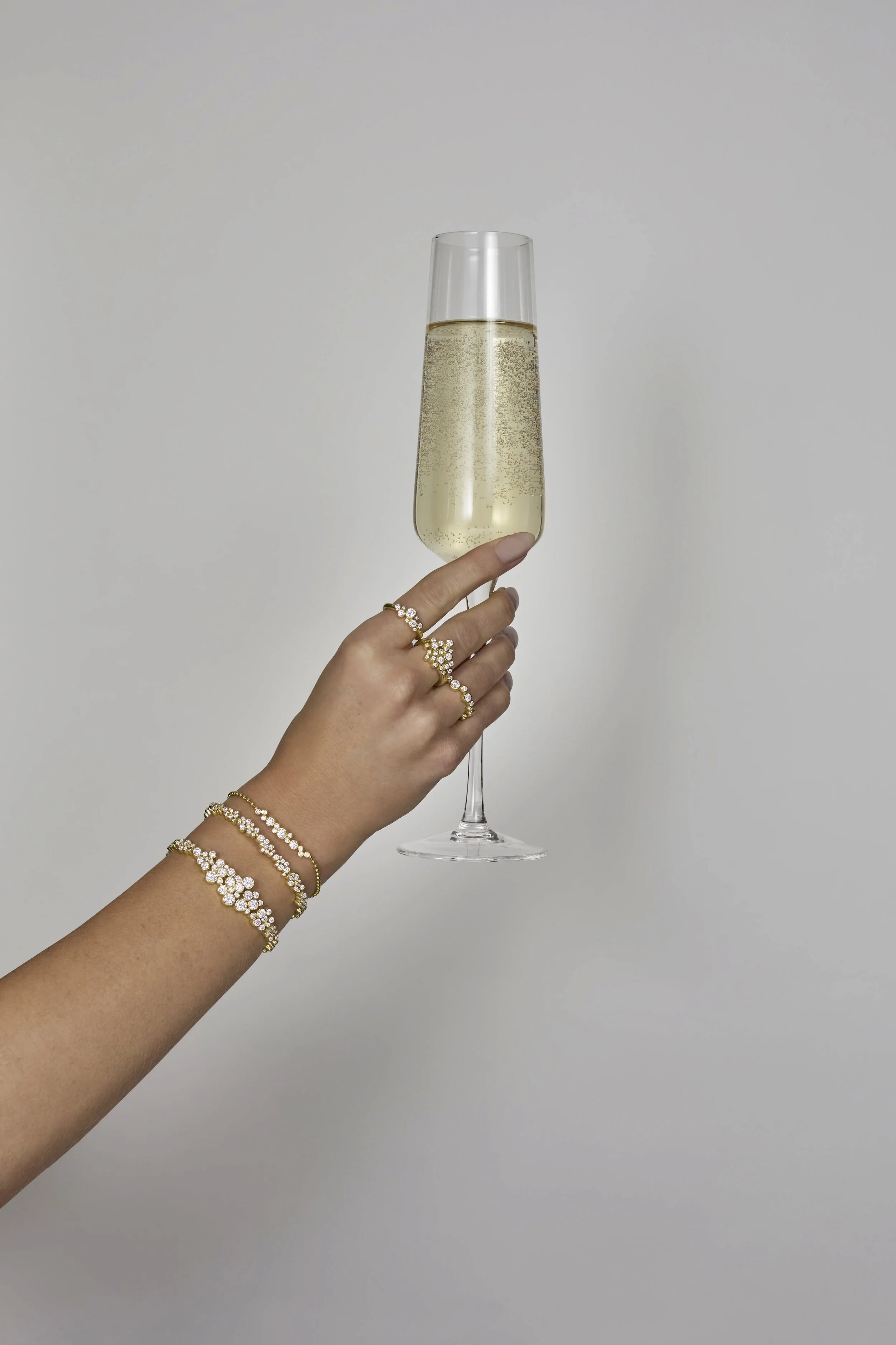 A hand holding a champagne flute filled with champagne against a plain background, adorned with gold jewelry.