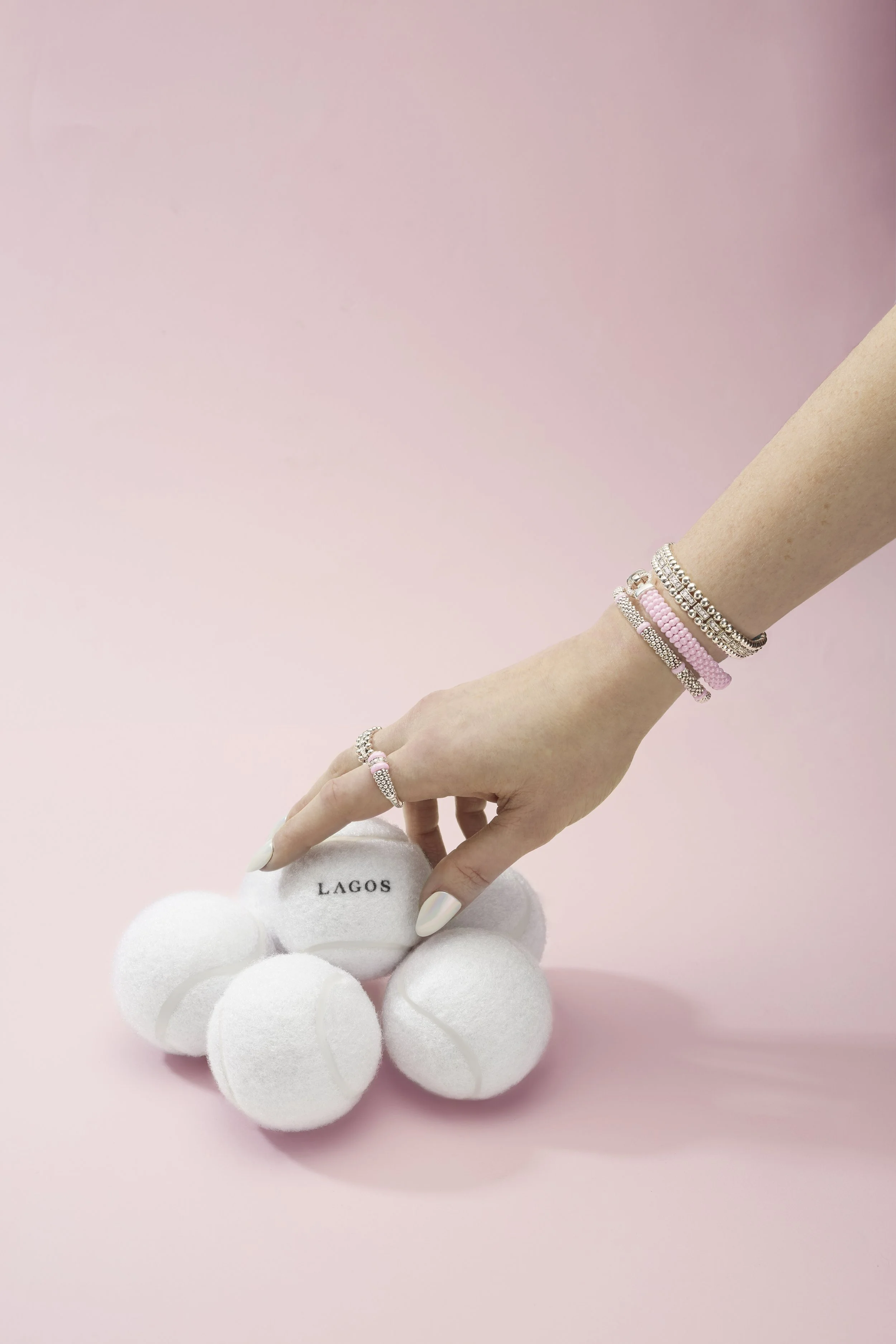 A hand with rings and bracelets is reaching for a white tennis ball with the word 'LAGOS' written on it, surrounded by additional tennis balls placed on a pink surface with a pink background.