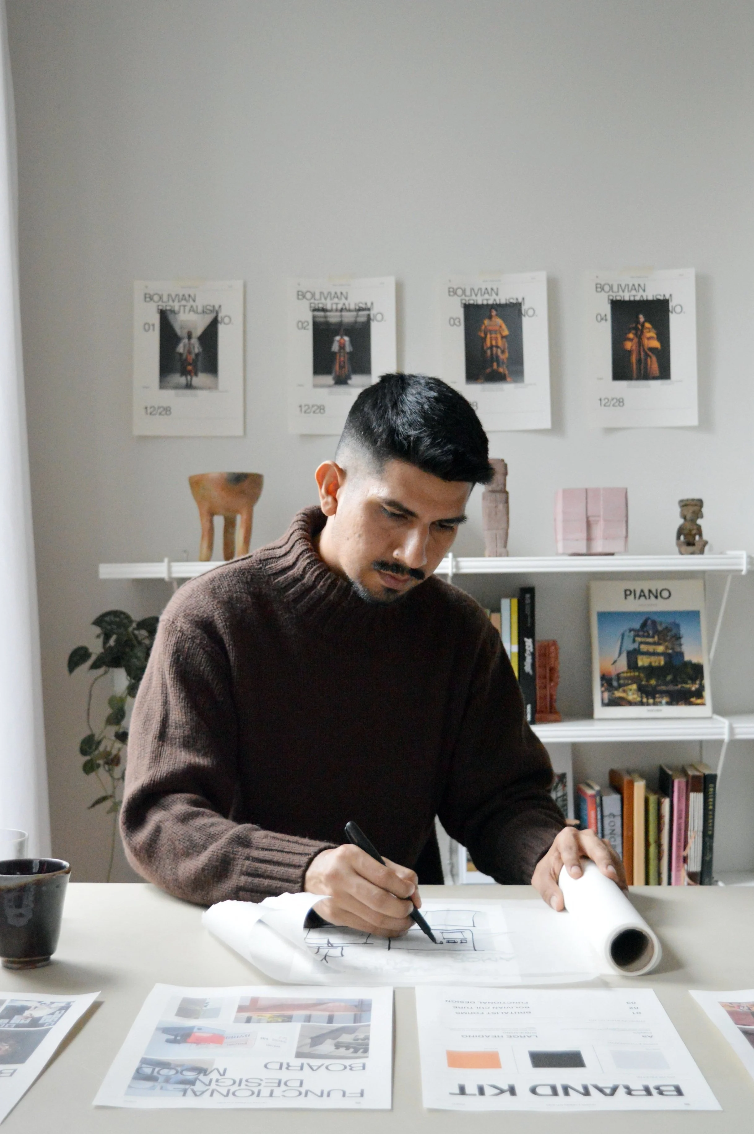 A man with dark hair and a mustache wears a brown sweater and is drawing plans at a white table. On the table are printed documents, a cup, and a rolled-up blueprint. Behind him is a white bookshelf with books, framed photos, and small sculptures. The wall above has four posters of clothing designs and a shelf with a plant and decorative items.