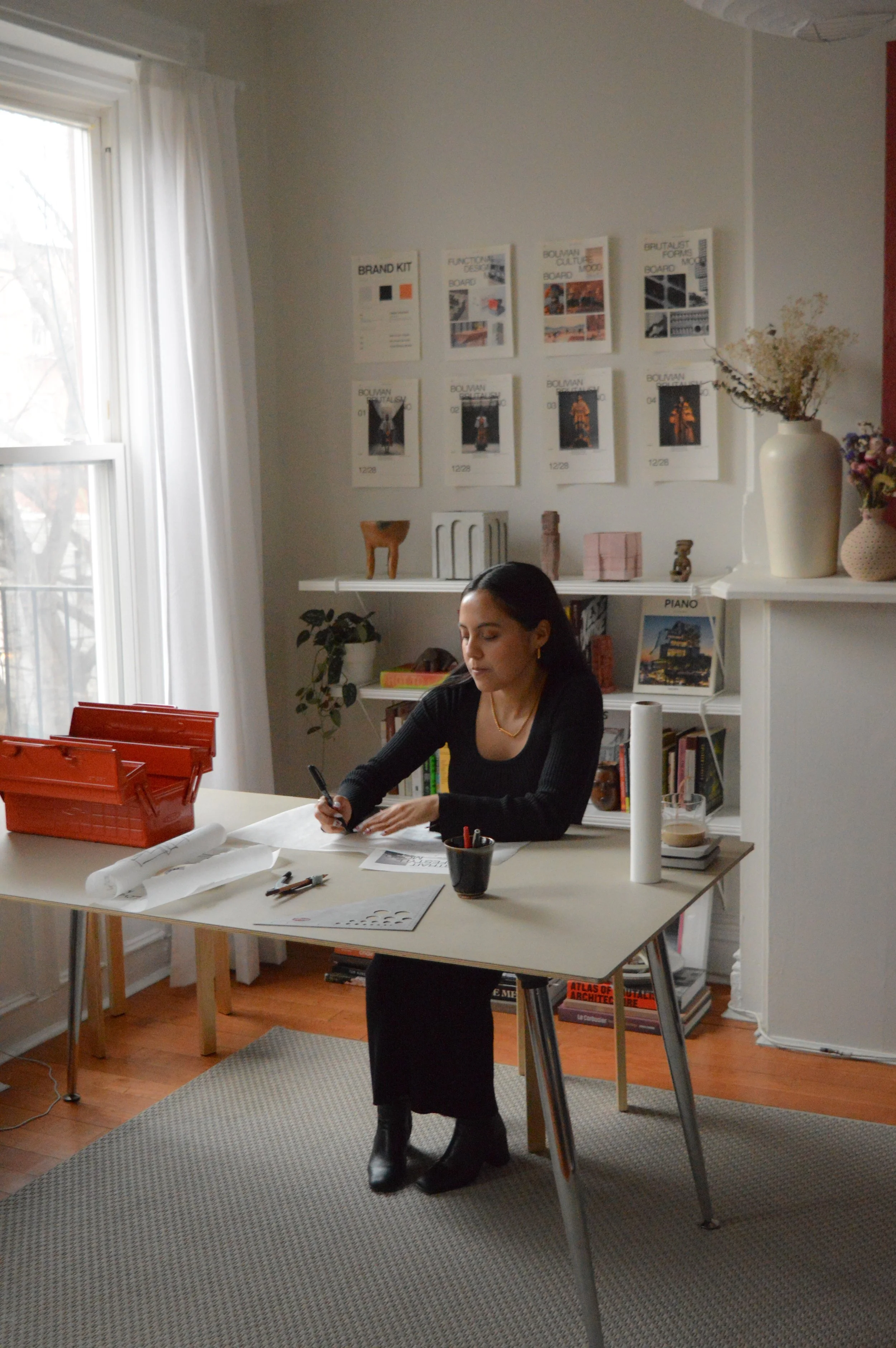 A woman sitting at a desk working on papers in a bright room with a large window, white curtains, and a bookshelf filled with books and decorative items in the background.