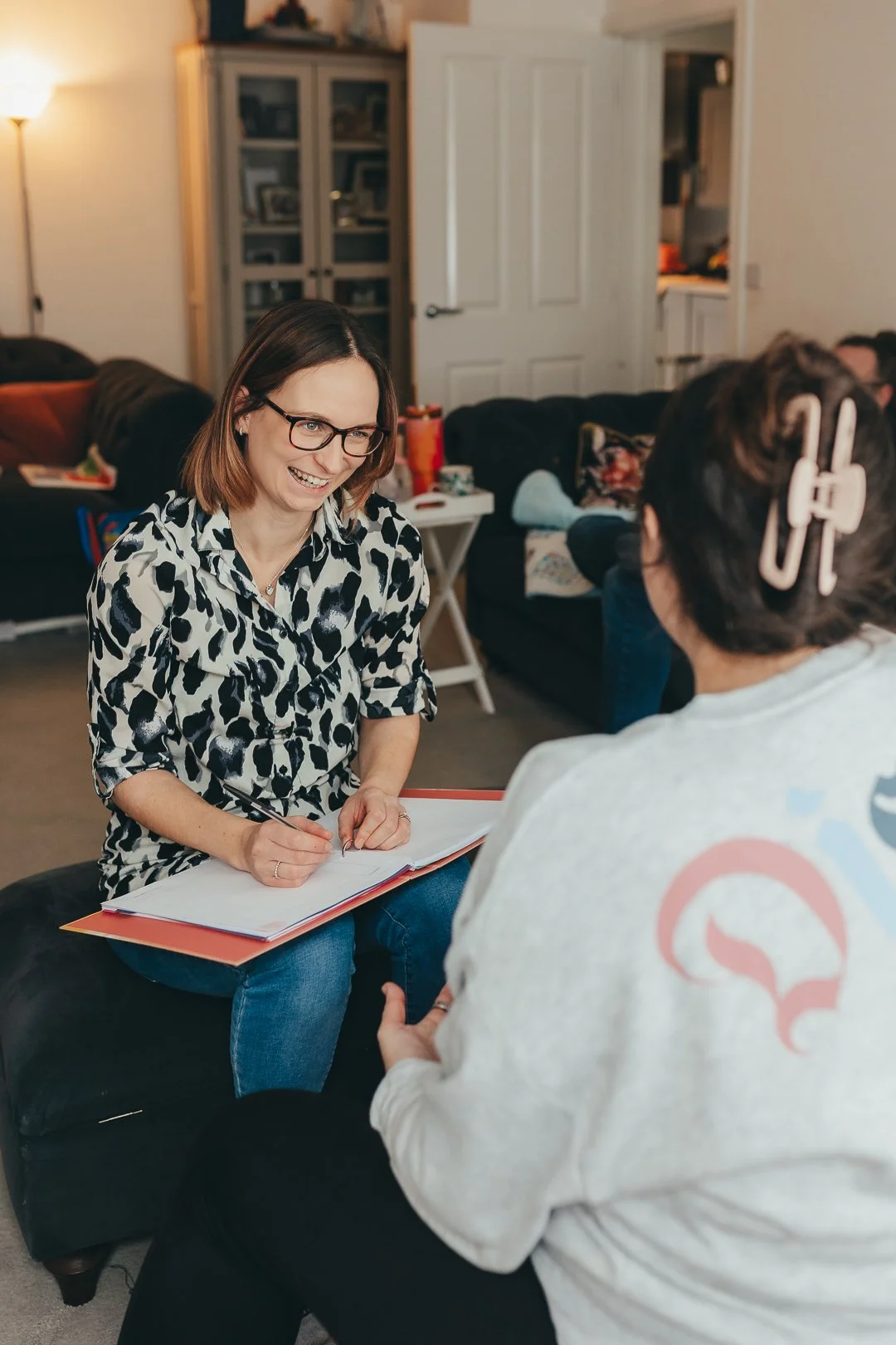 A woman with shoulder-length brown hair, glasses, and wearing a black and white animal print shirt, is smiling while sitting on a black sofa and writing in a large pink notebook. She is talking to a person with dark hair in a bun, wearing a light-colored sweatshirt with a graphic on the back, who is sitting across from her. The setting appears to be a cozy living room with a bookshelf and other people in the background.