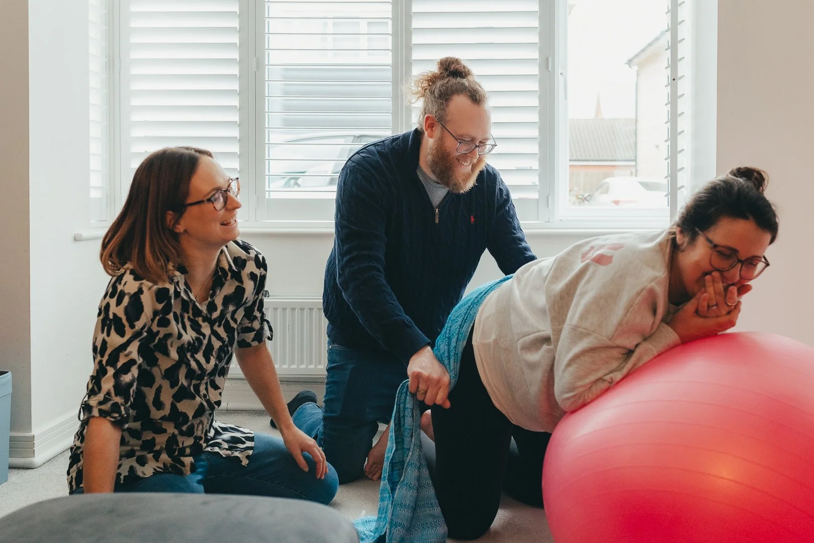 A woman is doing a hamstring stretch on an exercise ball while two people assist her in a lighthearted manner, indoors near a window with shutters.