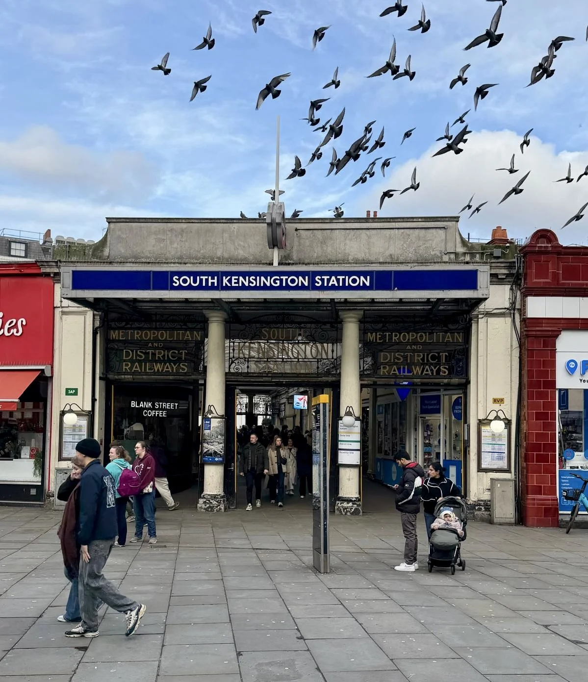 People walking and standing outside South Kensington Station in London, with pigeons flying above and a partly cloudy sky.