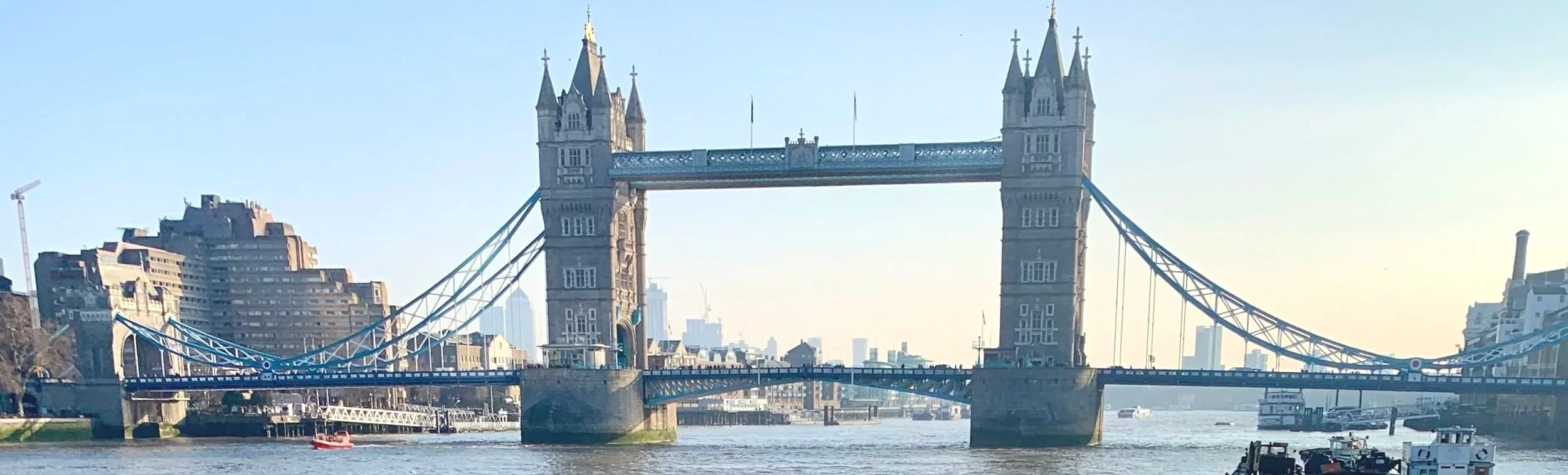 The Tower Bridge in London, seen from the river Thames with some boats passing underneath, and modern buildings in the background.