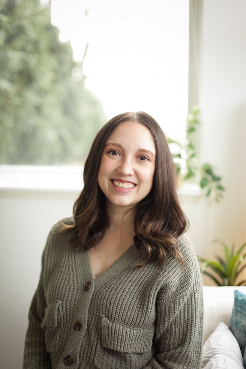 A woman with shoulder-length wavy brown hair and a beige knit cardigan smiling at the camera, sitting in a bright room with a window and greenery in the background.