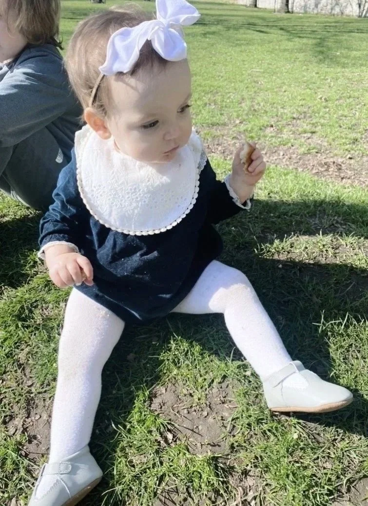 A young girl sitting on the grass outdoors, wearing a navy blue dress, white tights, white shoes, and a white bib with a lace trim, holding a small object in her hand, with a white bow headband on her head, and earing Lillie grey shoes by Nancy D's