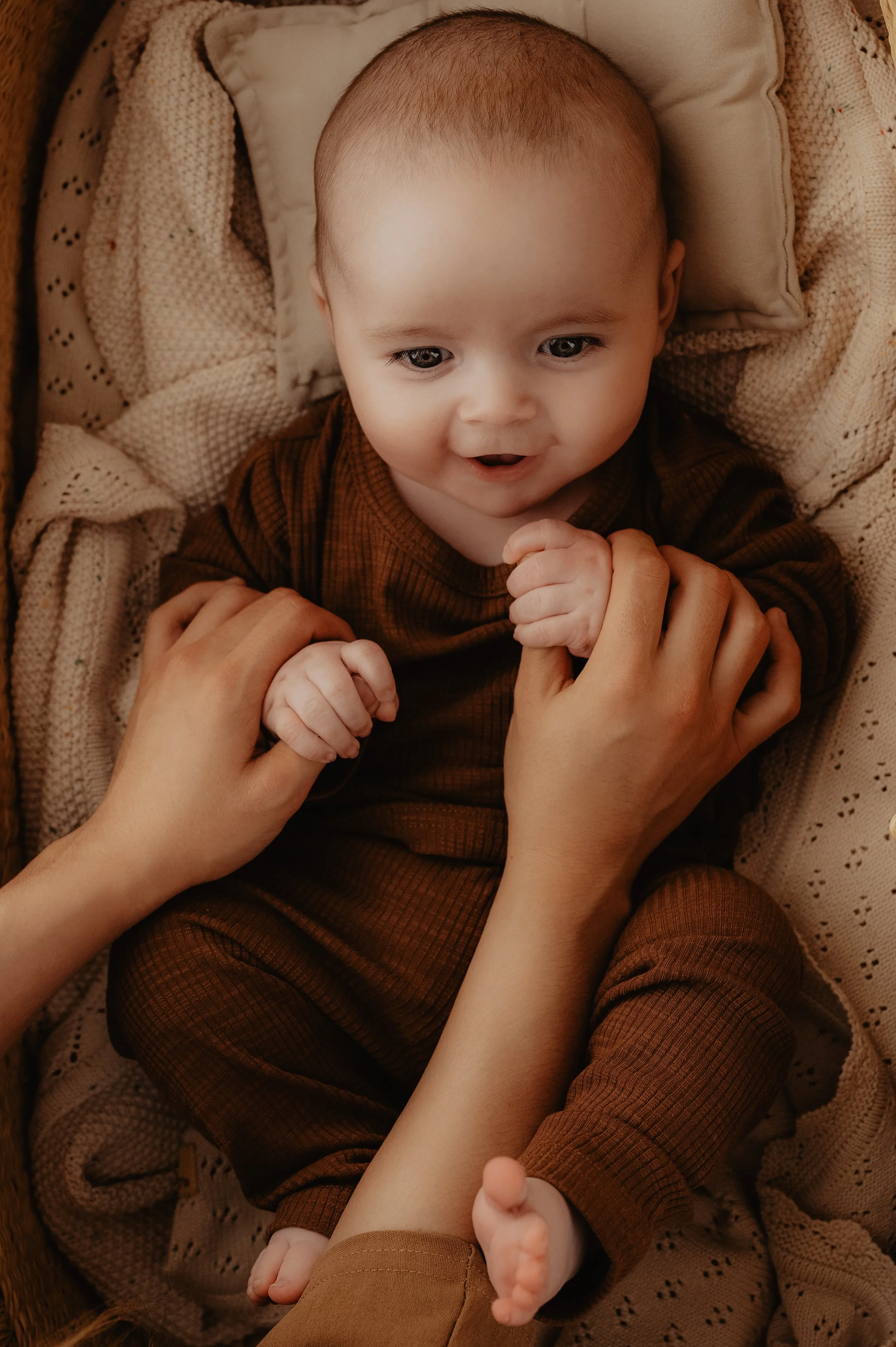A baby lying in a crib, holding hands with an adult, smiling and looking up wearing a Lennie outfit by Nancy D's in the colour brown