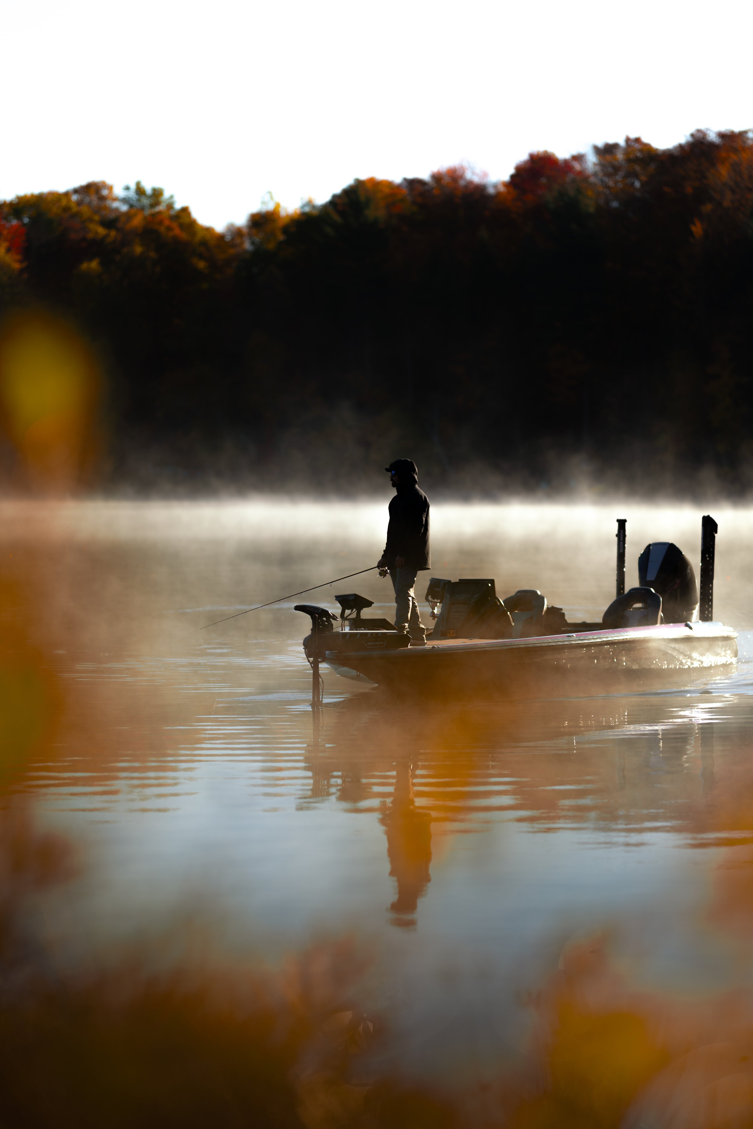 A person fishing on a boat on a foggy river during autumn.