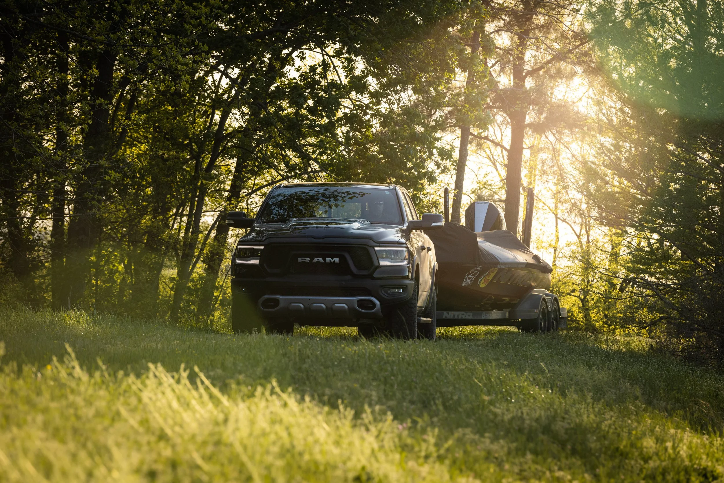 A black RAM pickup truck towing a speedboat on a trailer through a grassy and wooded area during sunset.