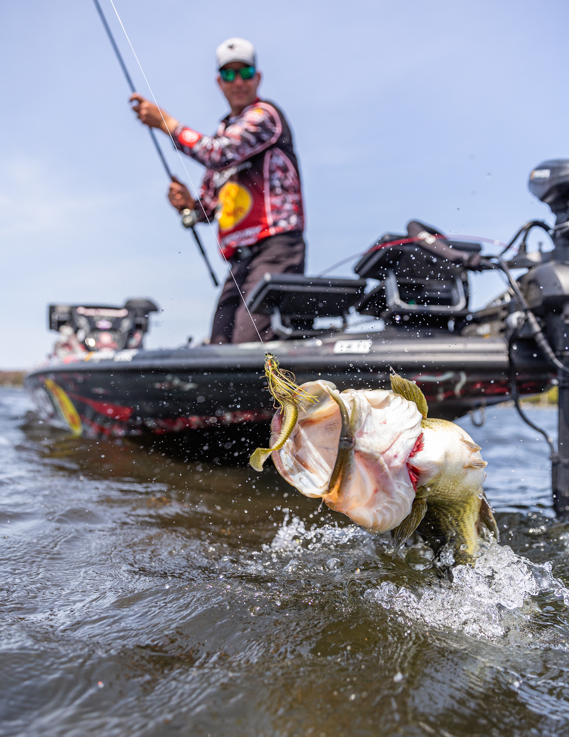 A person in sunglasses, a cap, and a fishing jersey on a boat, holding a fishing rod, has just caught a large fish, which is splashing water as it is being reeled in.