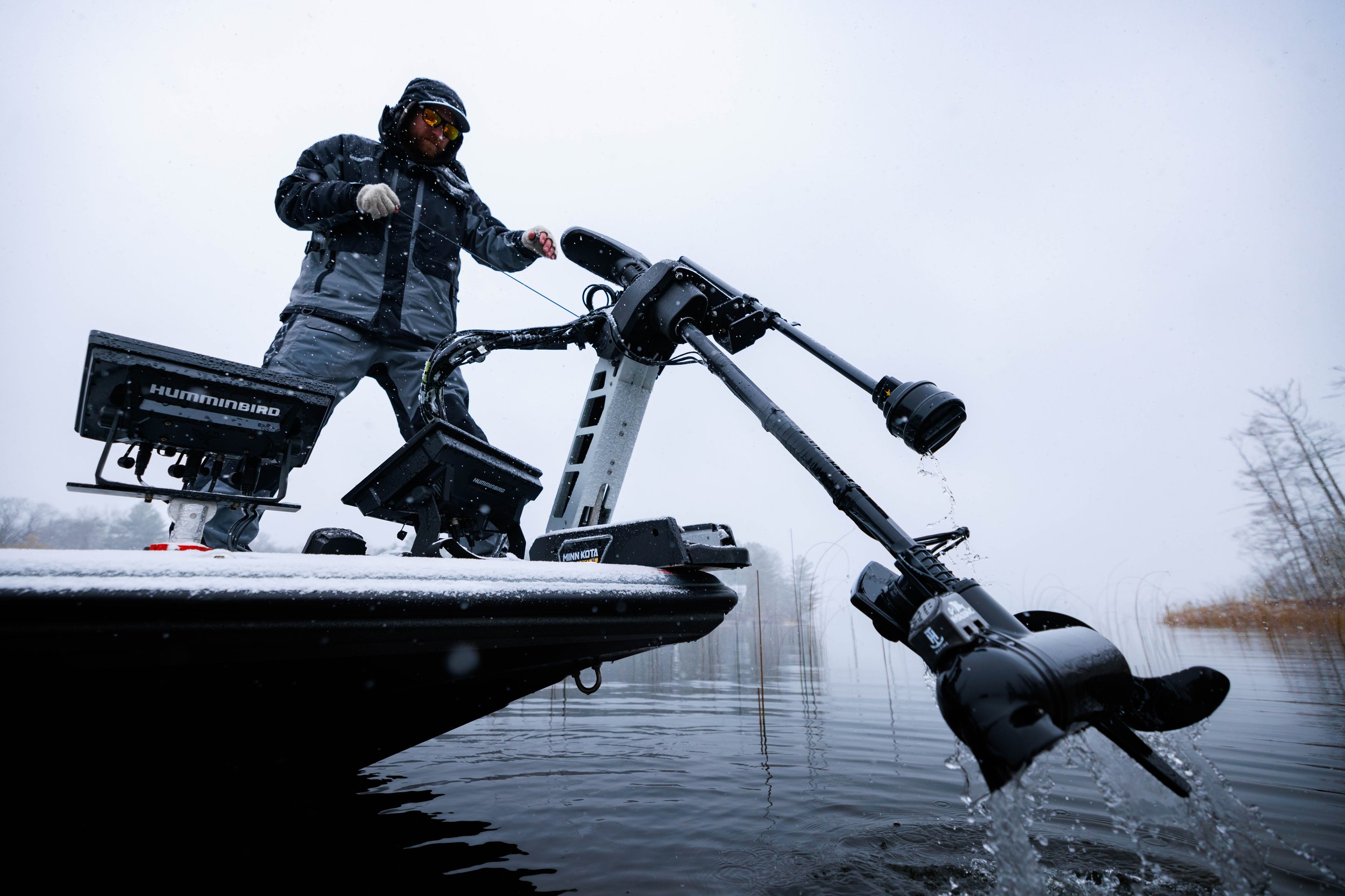 Person in winter gear operating a trolling motor on a boat in a snowy outdoor setting.