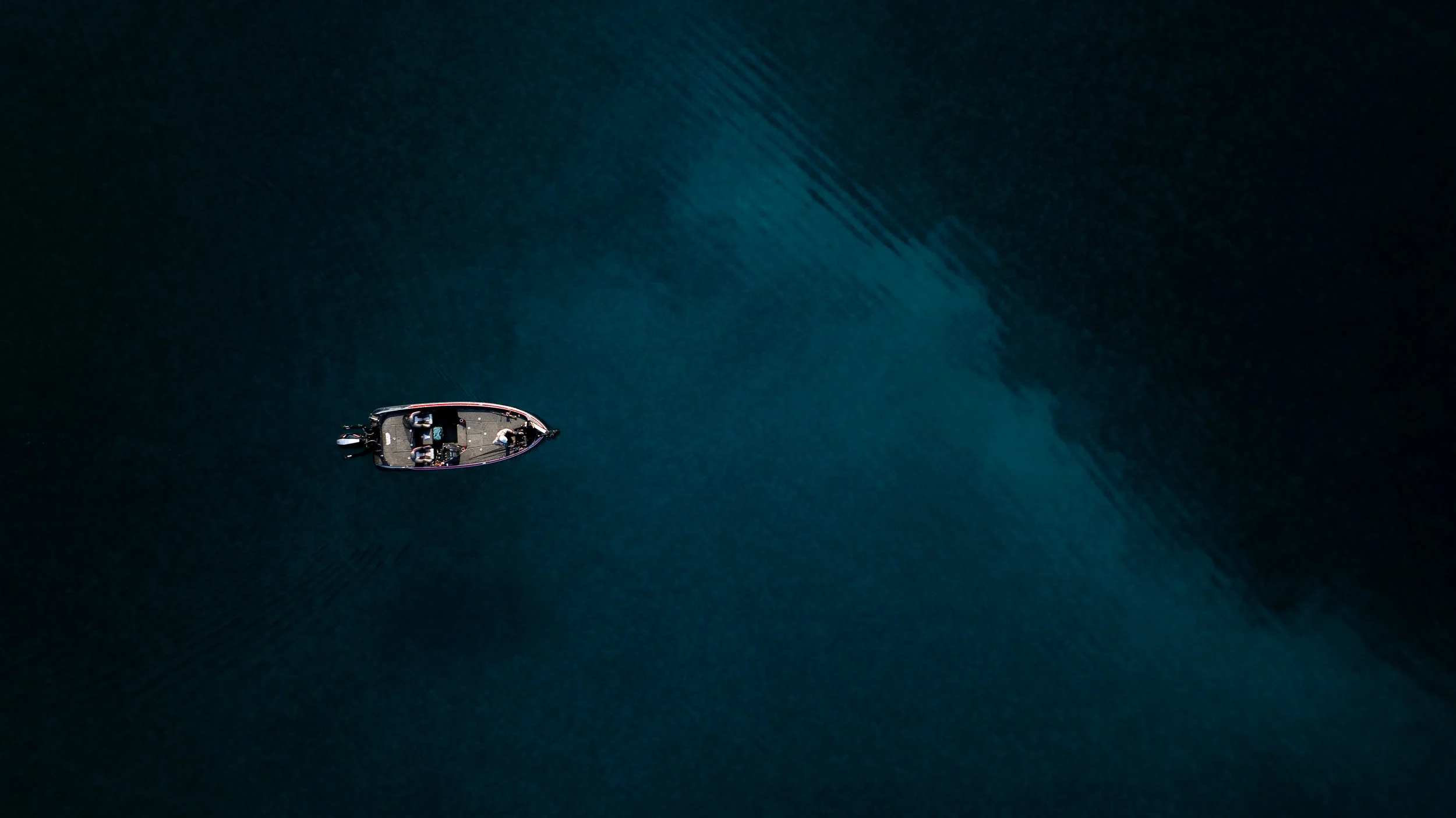 Aerial view of a small boat with two people, moving through dark blue water, leaving a wake behind.