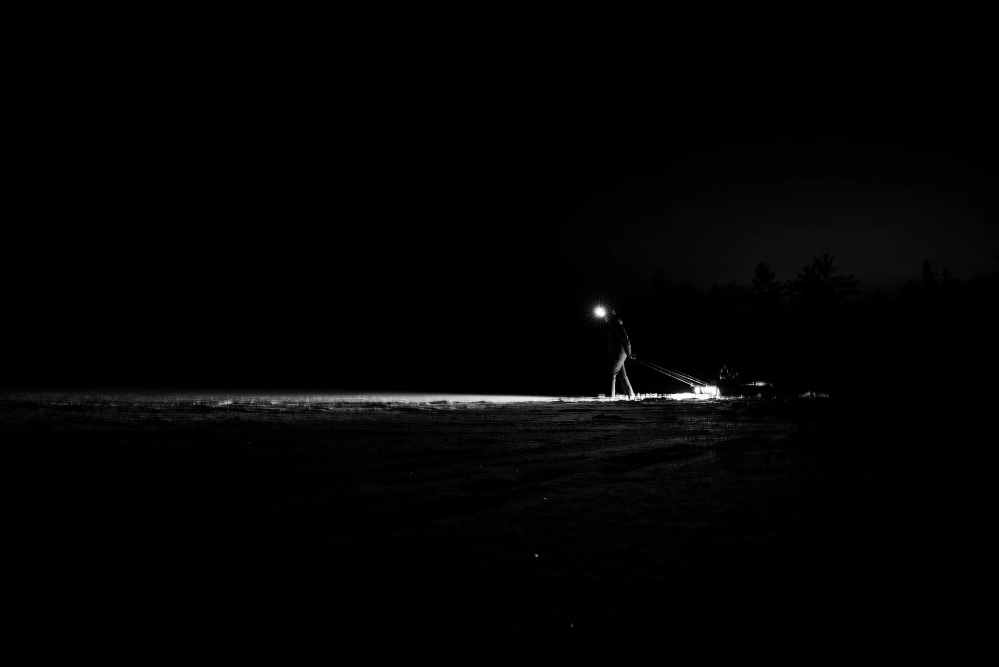 A person walking on a dark, snowy landscape at night, wearing a headlamp and pulling a sled with supplies, with a forest silhouette in the background.
