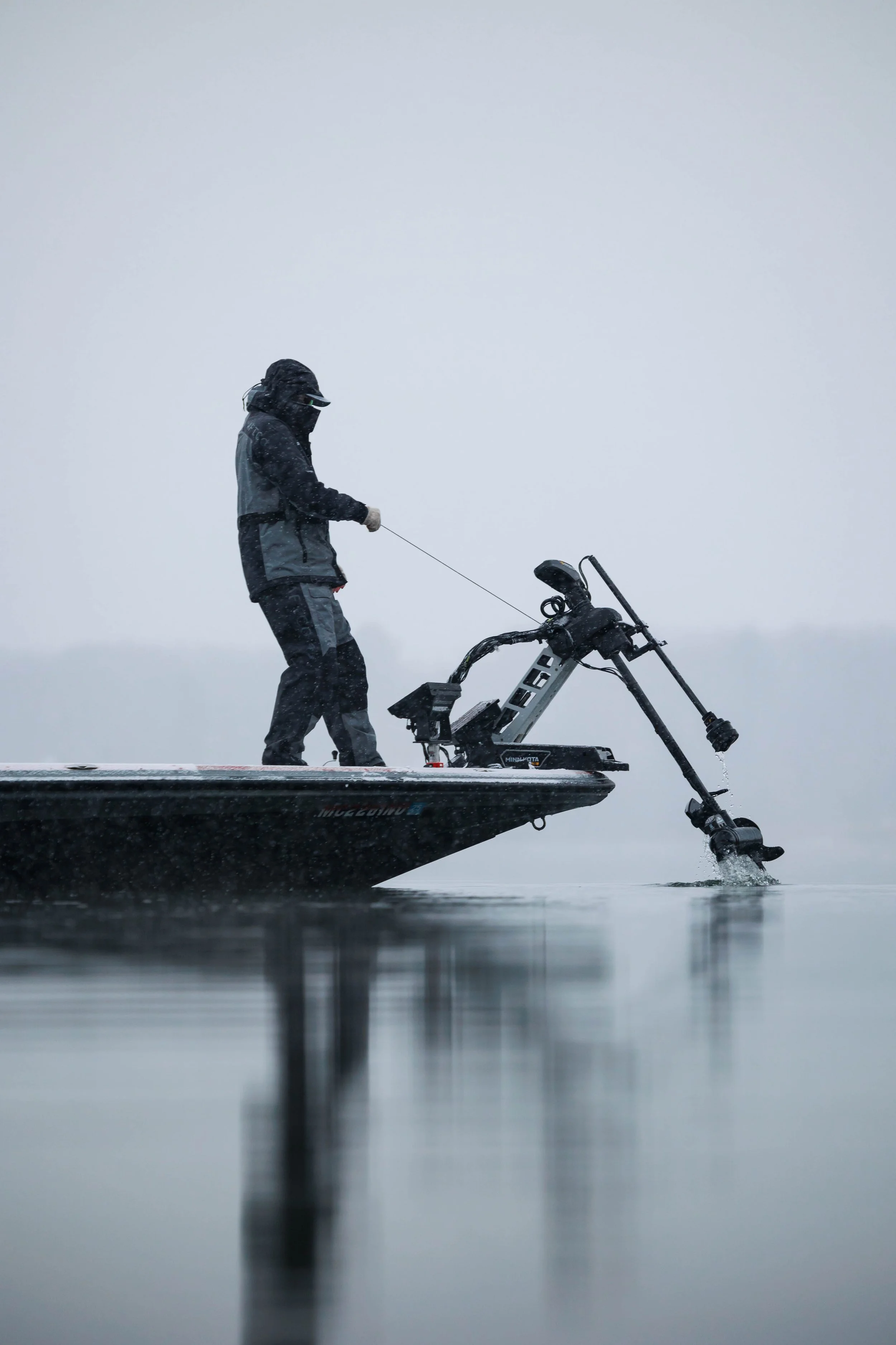 Person fishing from a boat on a calm, foggy lake with gray sky.