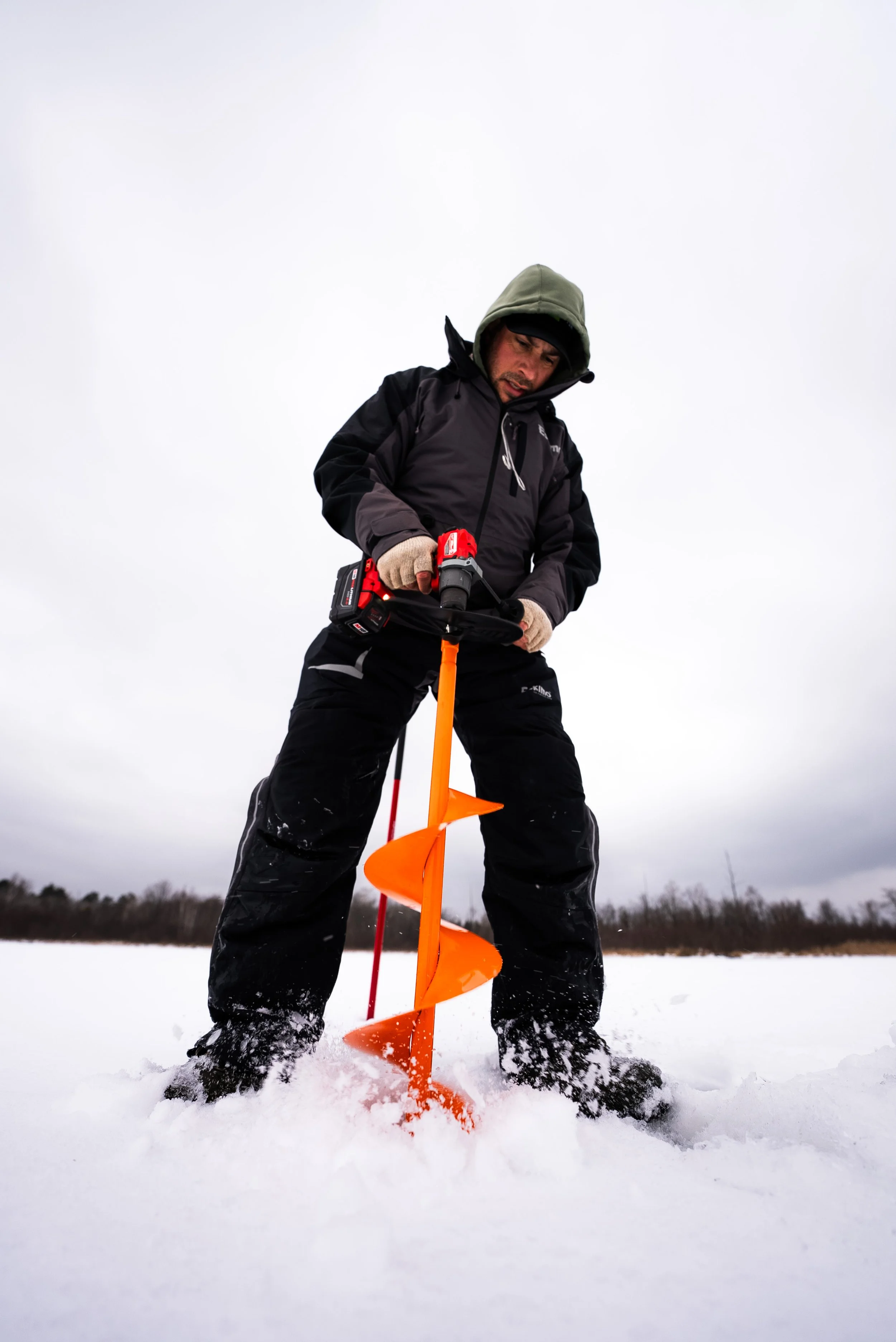 Man in winter clothing using an orange ice auger on a snowy landscape overcast sky.