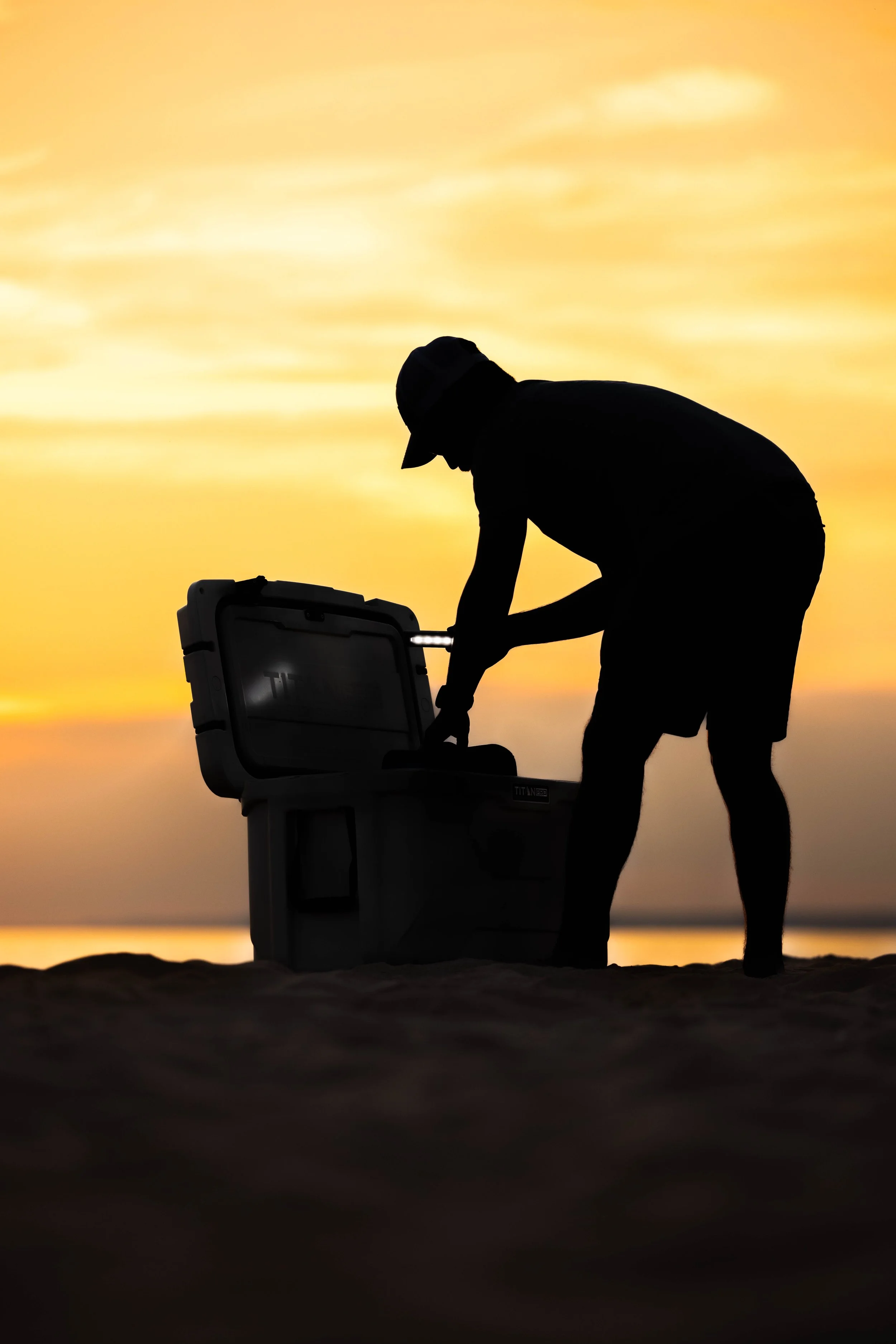 Silhouette of a man at the beach opening a cooler during sunset.