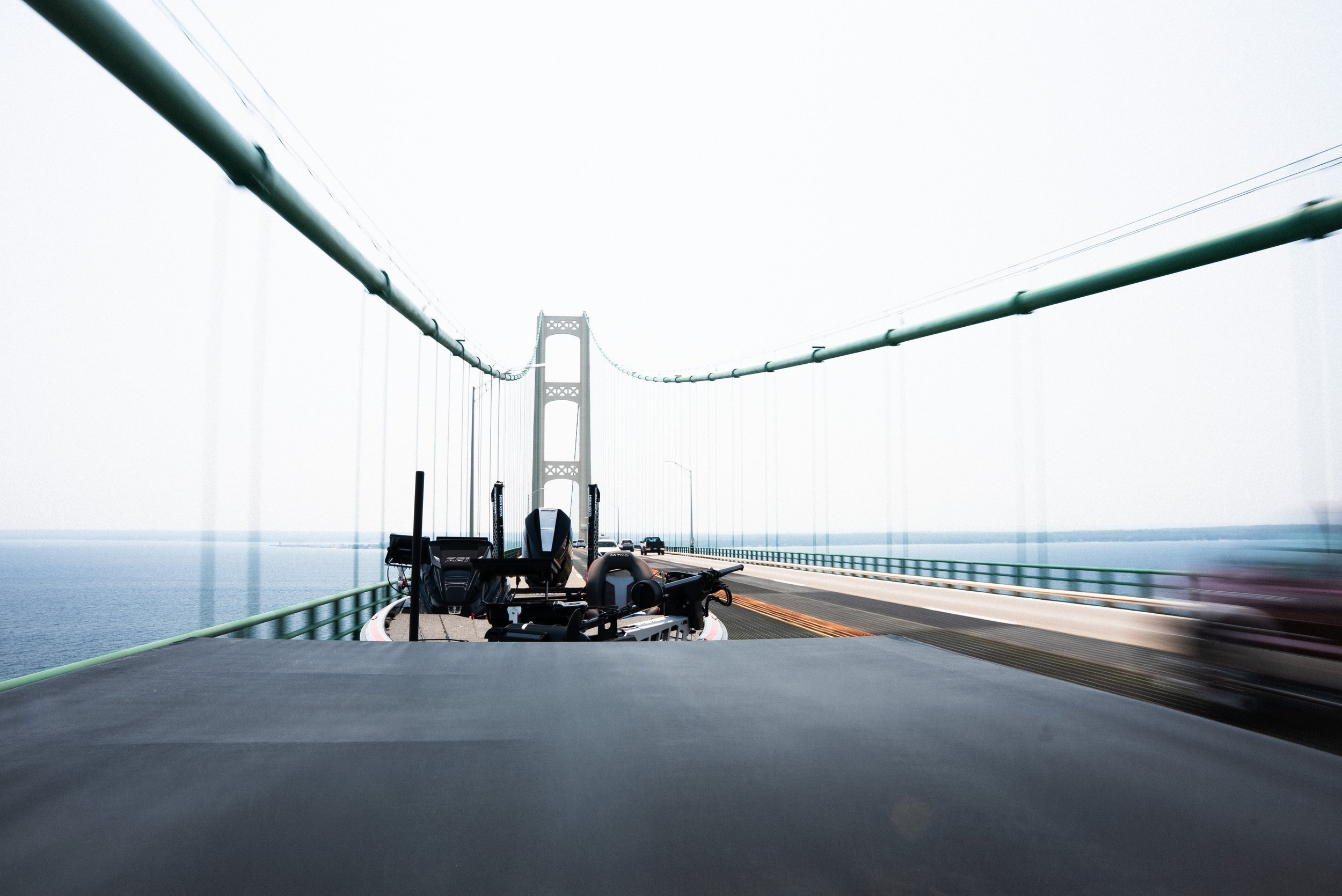 View from a vehicle on a bridge over water, showing equipment in the front and the bridge's structure with cables and towers in the distance.