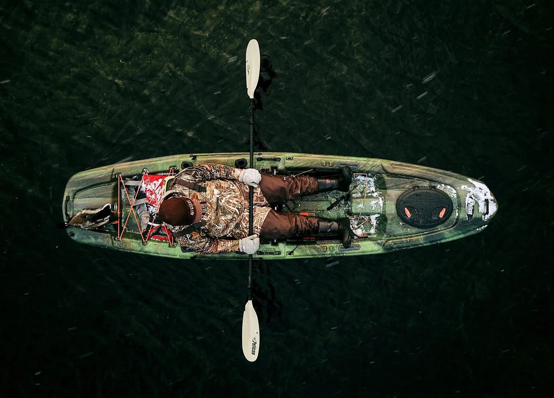 Top-down view of a person kayaking on dark water, wearing camouflage jacket, brown pants, and a beanie, holding paddles.