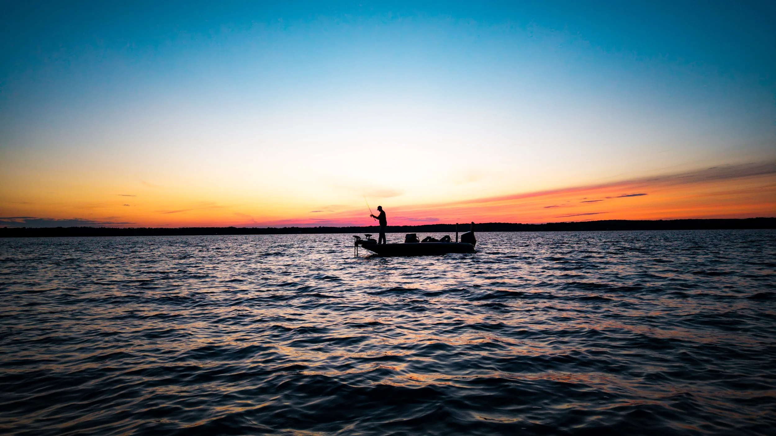 Silhouette of a person fishing on a boat during sunset on a calm lake.