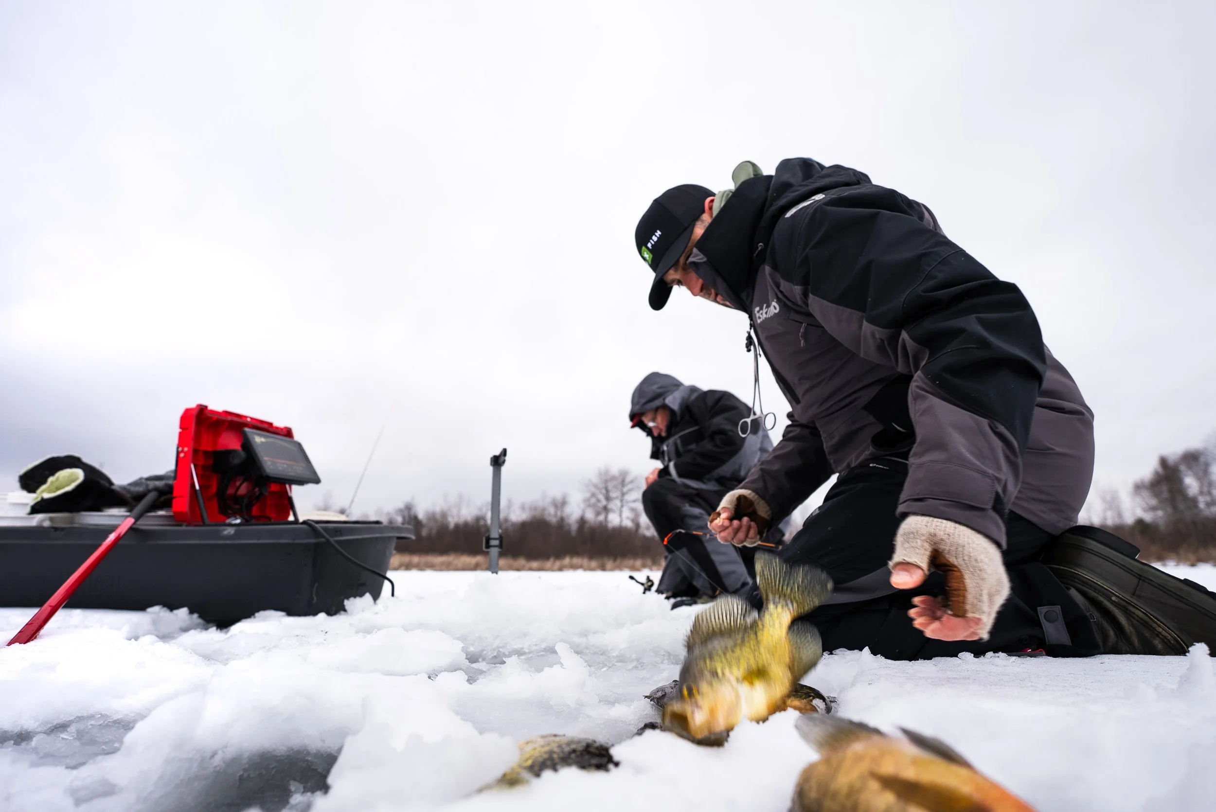 Two people ice fishing on a snow-covered lake, one holding a fish, with ice fishing equipment nearby.