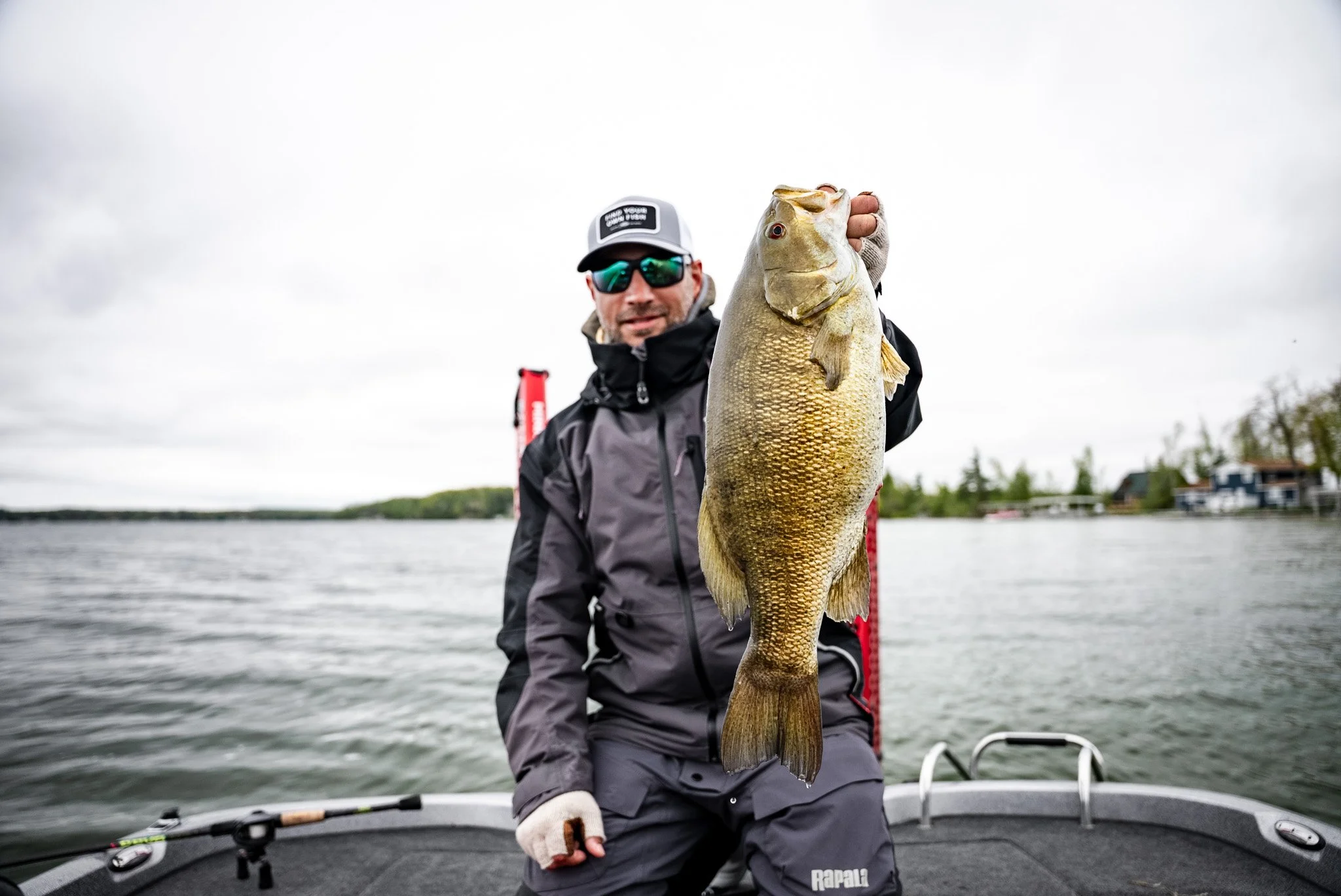 Man in black fishing gear holding a large fish on a boat with water and trees in the background.