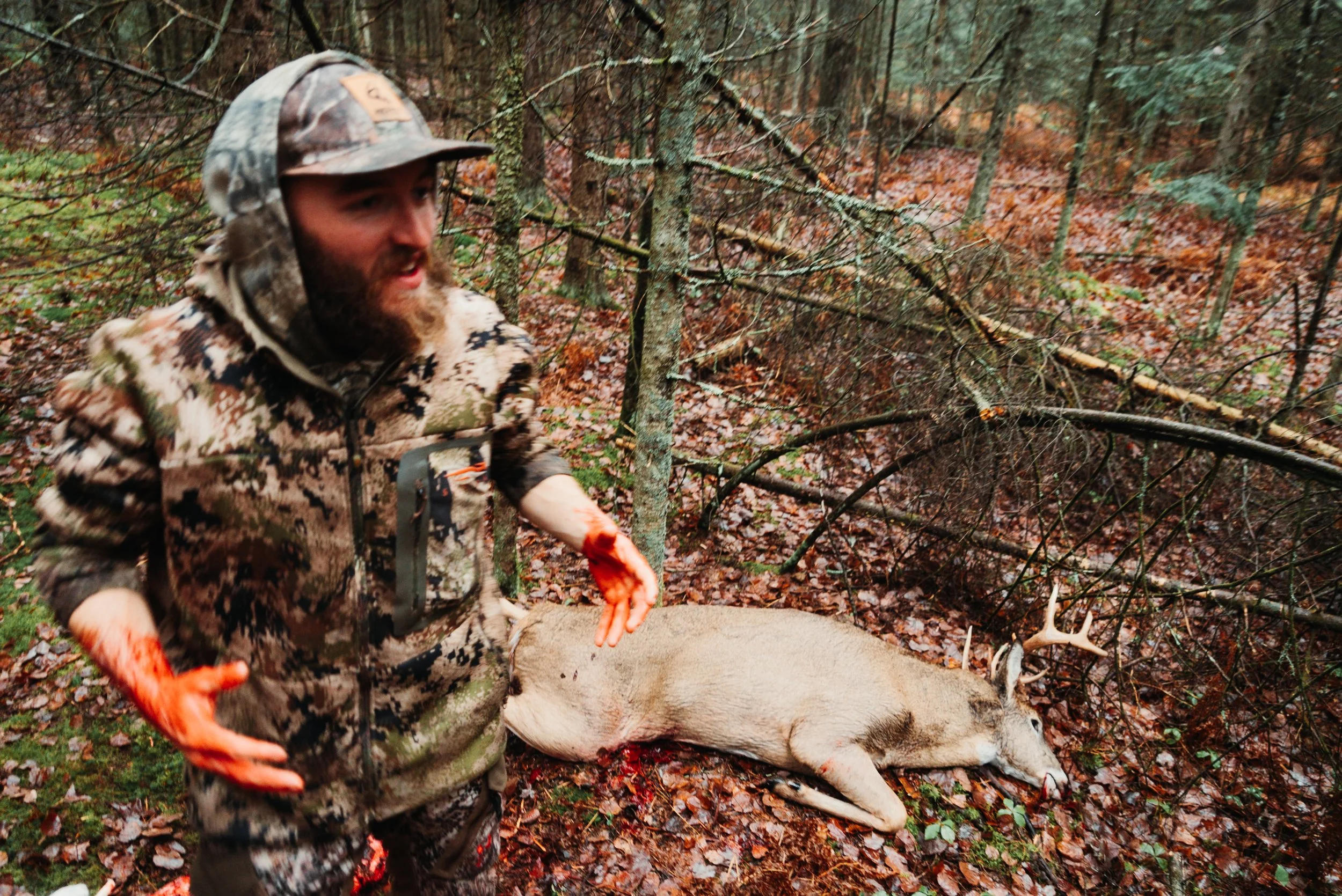 A hunter wearing camouflage clothing and orange gloves stands in a forest near a dead deer laying on the ground, with fallen leaves and trees in the background.