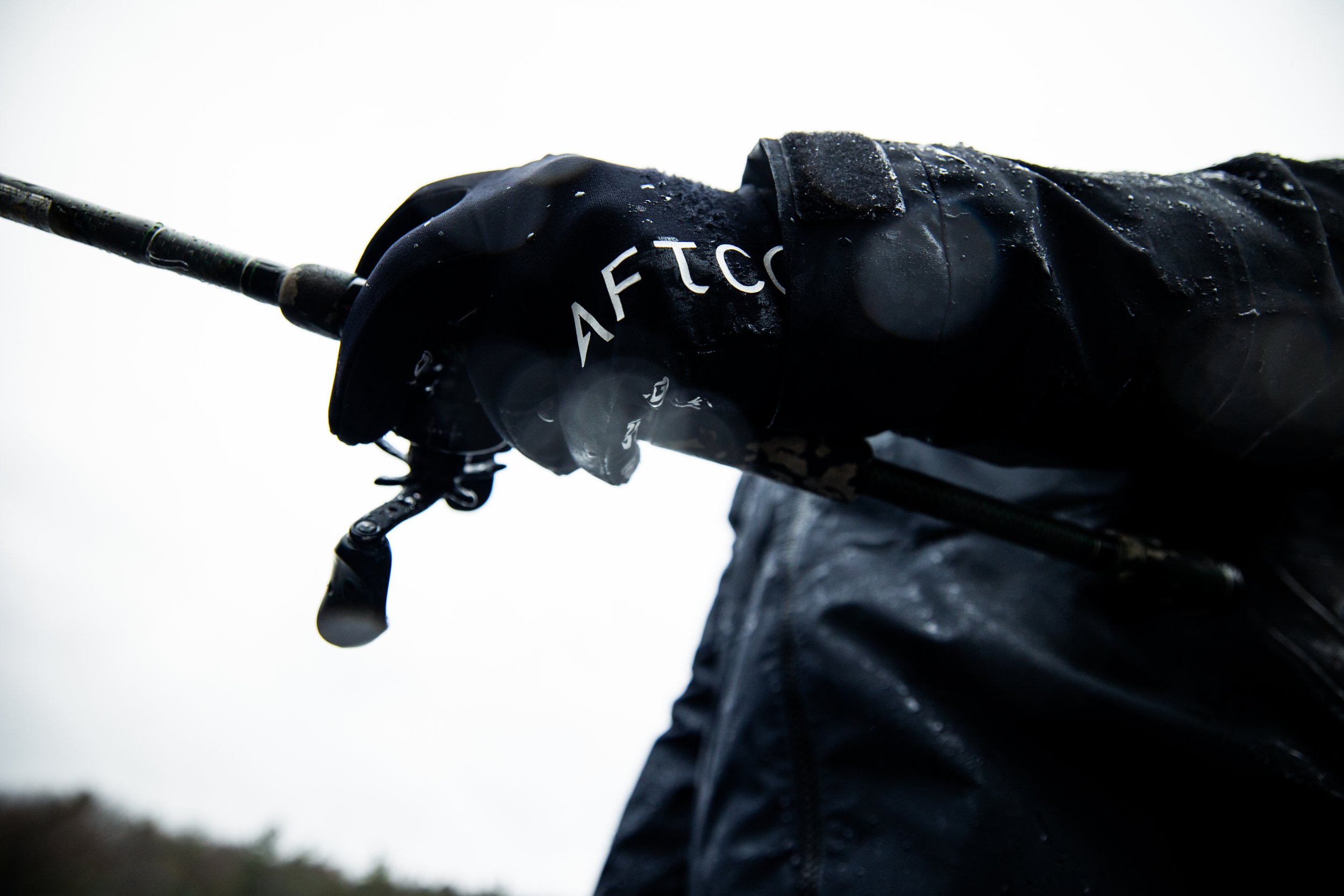 Close-up of a person's arm wearing a black waterproof jacket holding a fishing rod, with raindrops on the jacket and overcast sky in the background.