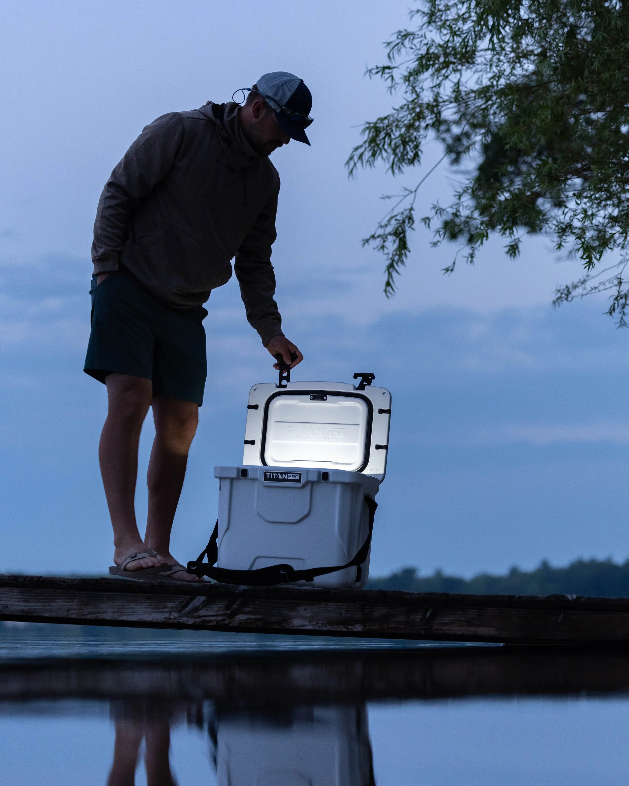 A man in a hoodie, shorts, and flip-flops standing on a dock at dusk, opening a white cooler labeled 'TITAN PRO' with a background of water and a cloudy sky.