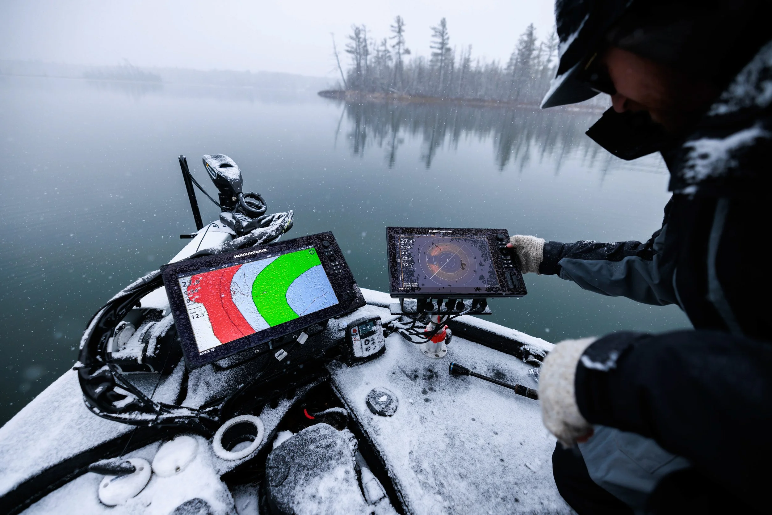 Person on a snow-covered boat using electronic navigation equipment on a frozen lake with a forested shoreline and overcast sky in the background.
