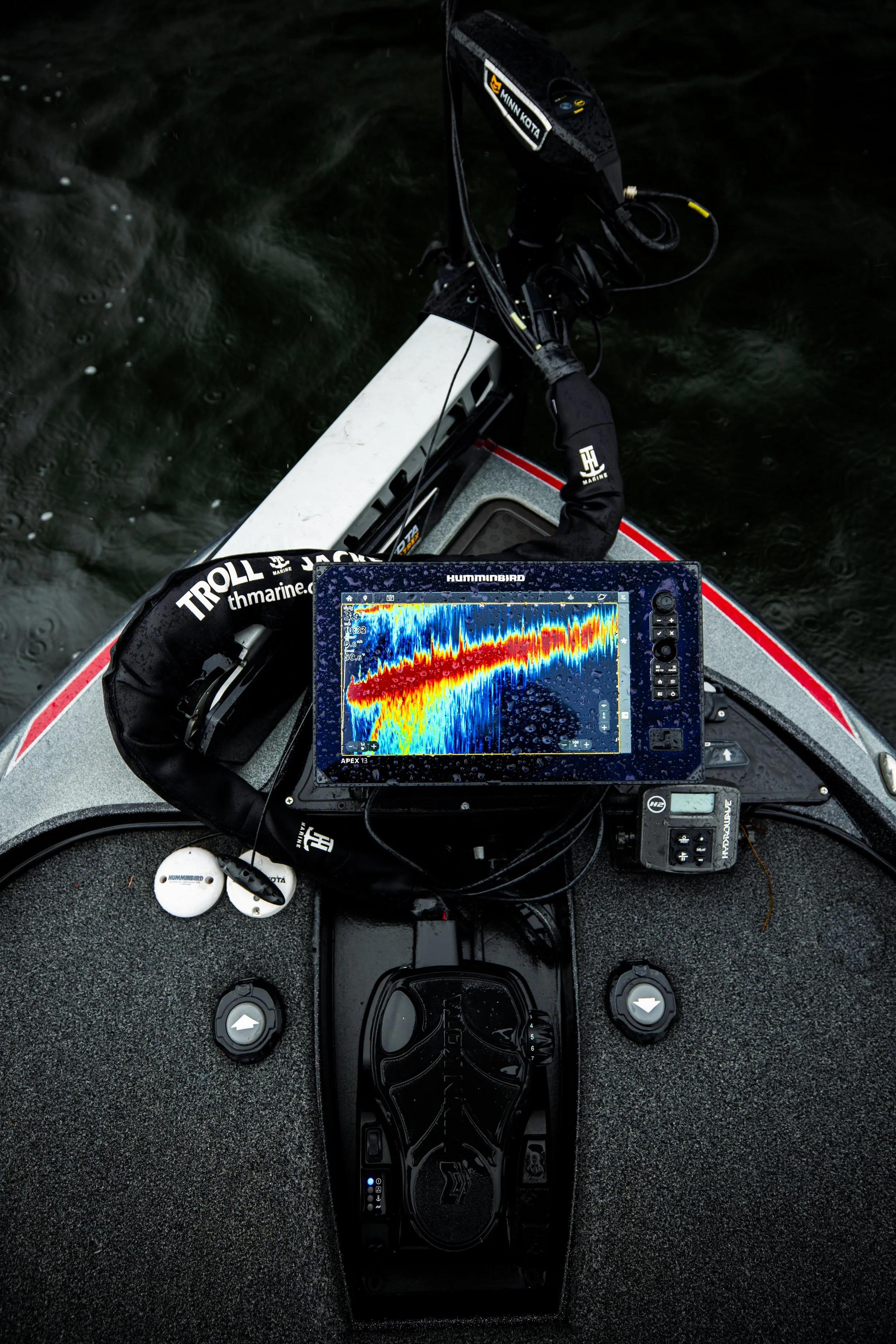 A fishing boat with a fish finder displaying colorful sonar images, a trolling motor, and various fishing equipment on a water surface.