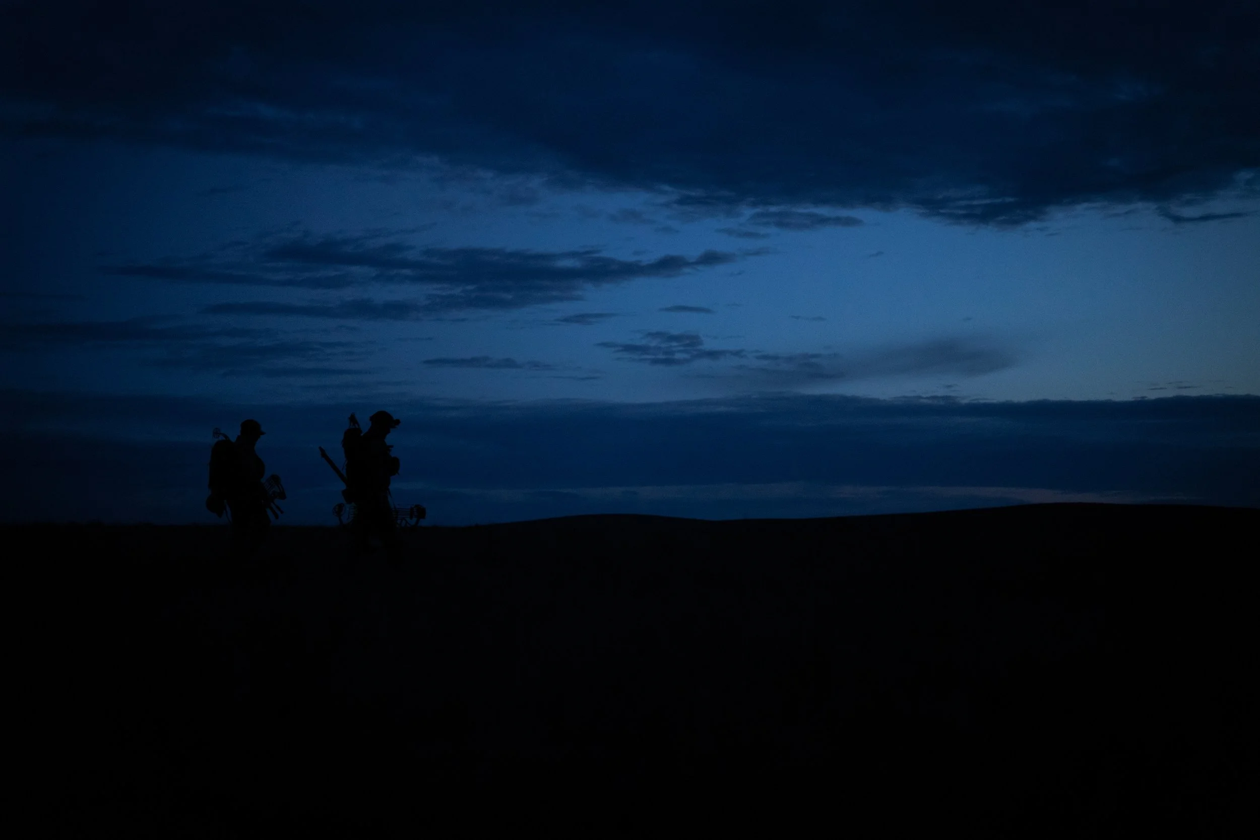 Silhouettes of two hikers walking across a dark landscape at dusk, with a cloudy sky overhead.