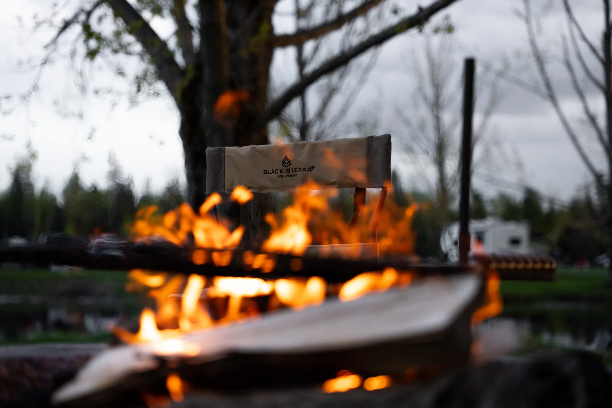 A campfire with burning flames and a beige bag labeled 'Black Sierra Equipment' in the background, outdoors with trees and a cloudy sky.