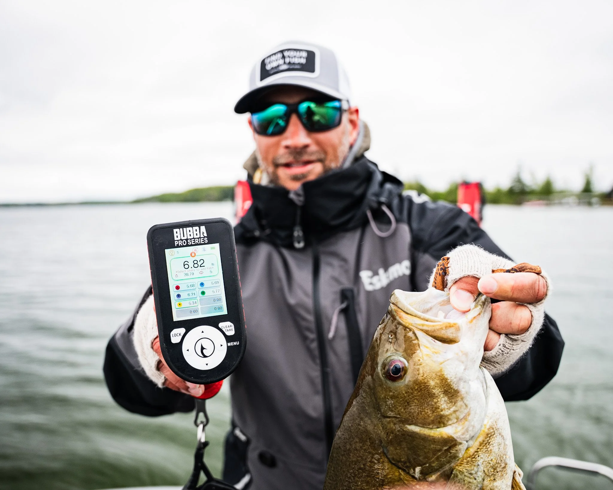 Man in outdoor gear holding a large fish in one hand and a fish finder device showing a 6.82-pound catch in the other, on a boat on a body of water.