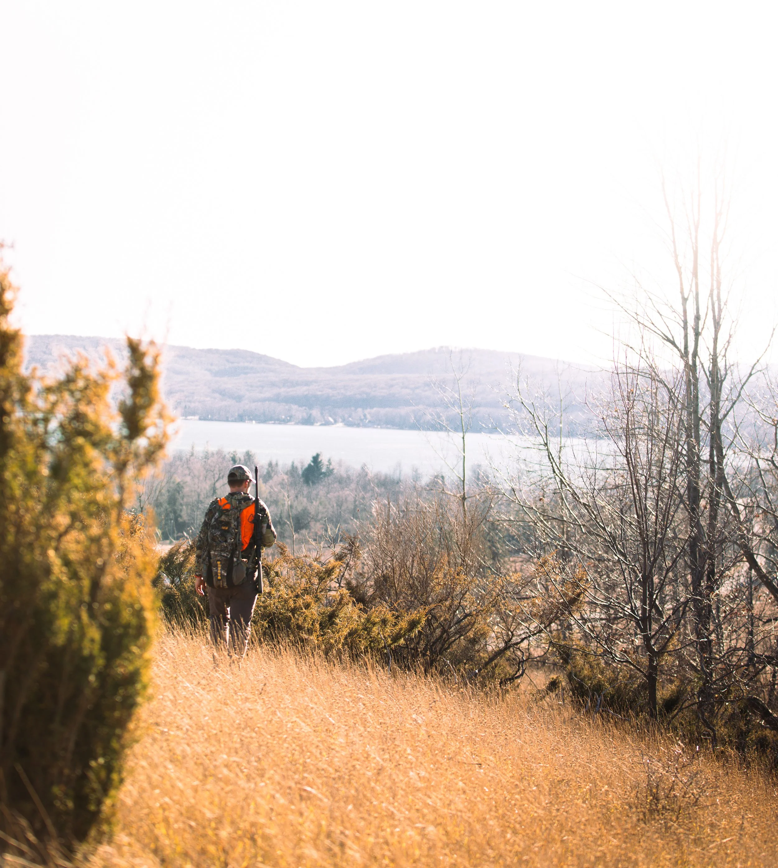 A hiker walking along a grassy trail through a nature landscape with trees and distant hills under a bright sky.