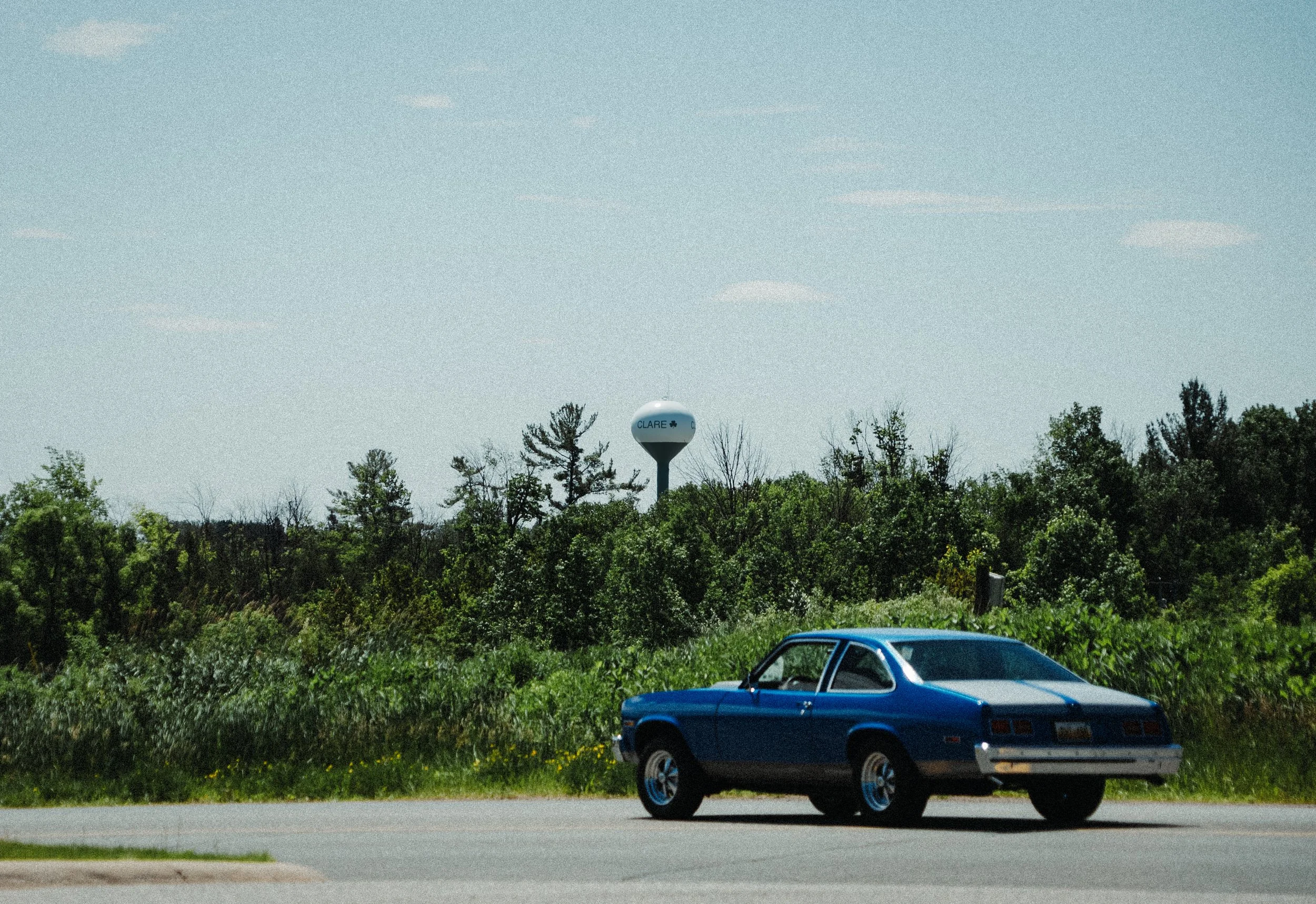 A vintage blue car parked on a roadside with green trees and bushes in the background and a water tower in the distance under a blue sky.