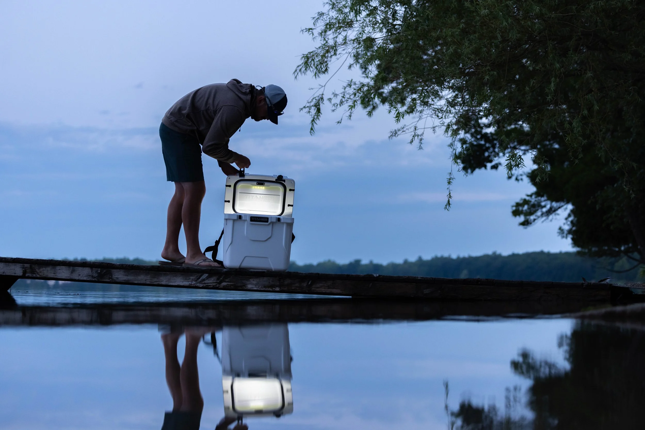 A person standing barefoot on a wooden dock, filling a white cooler with a flashlight inside, near a body of water at dusk with trees and cloudy sky in the background.