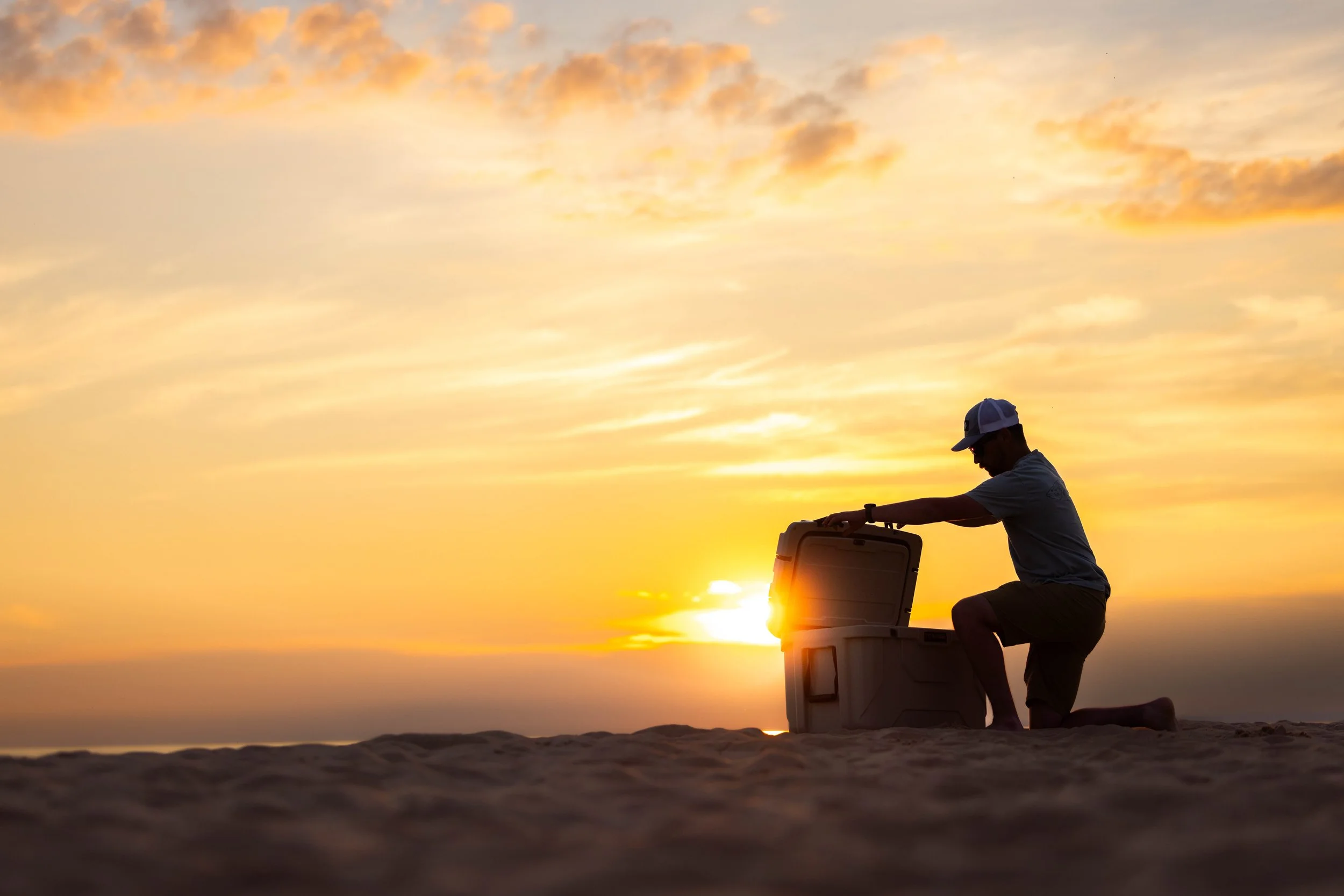 A man kneeling on the sand at sunset, opening a cooler, with the sun setting over the ocean in the background.