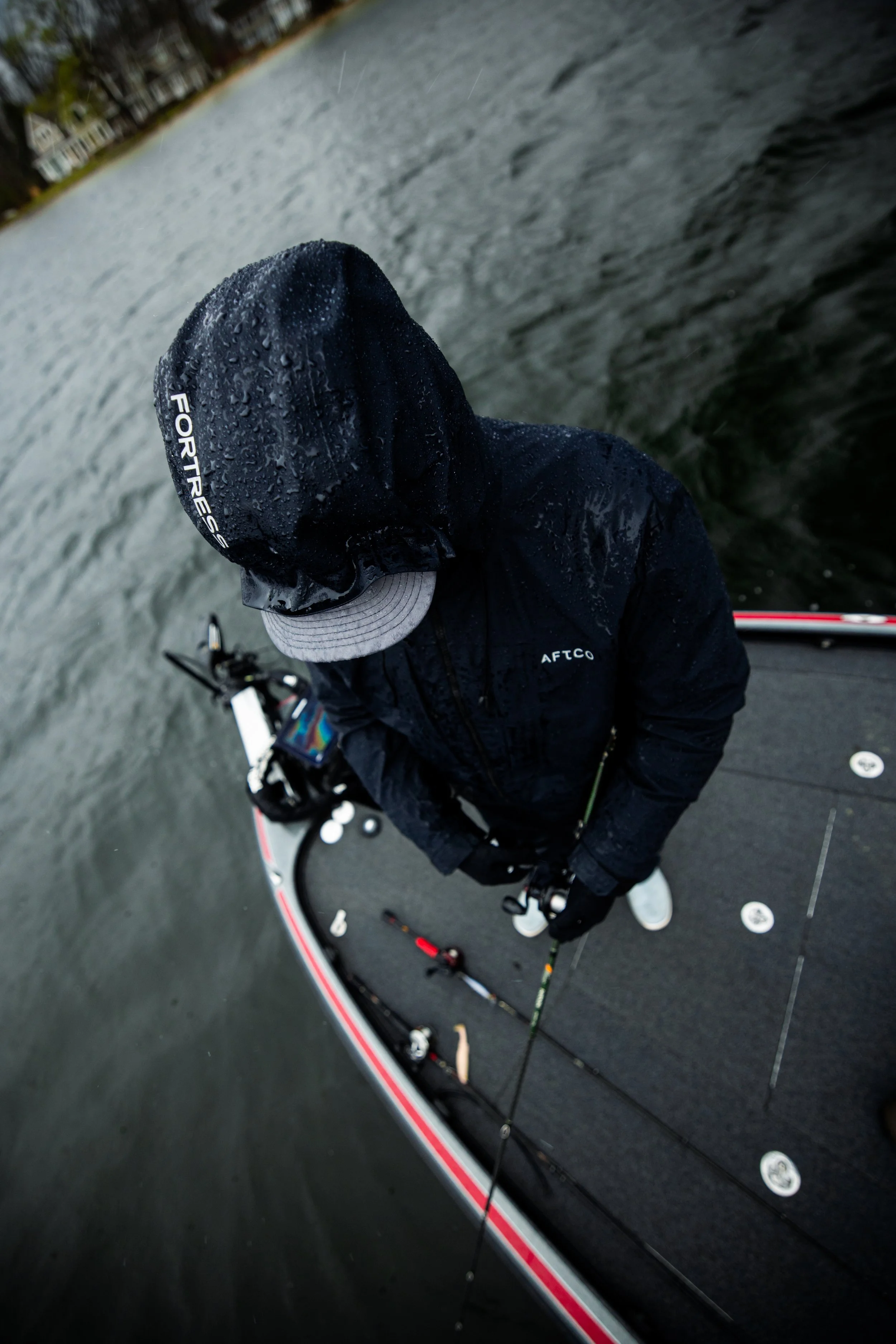 A person standing on a boat dressed in dark weather gear, including a hooded jacket and cap, holding a fishing rod on a rainy day.