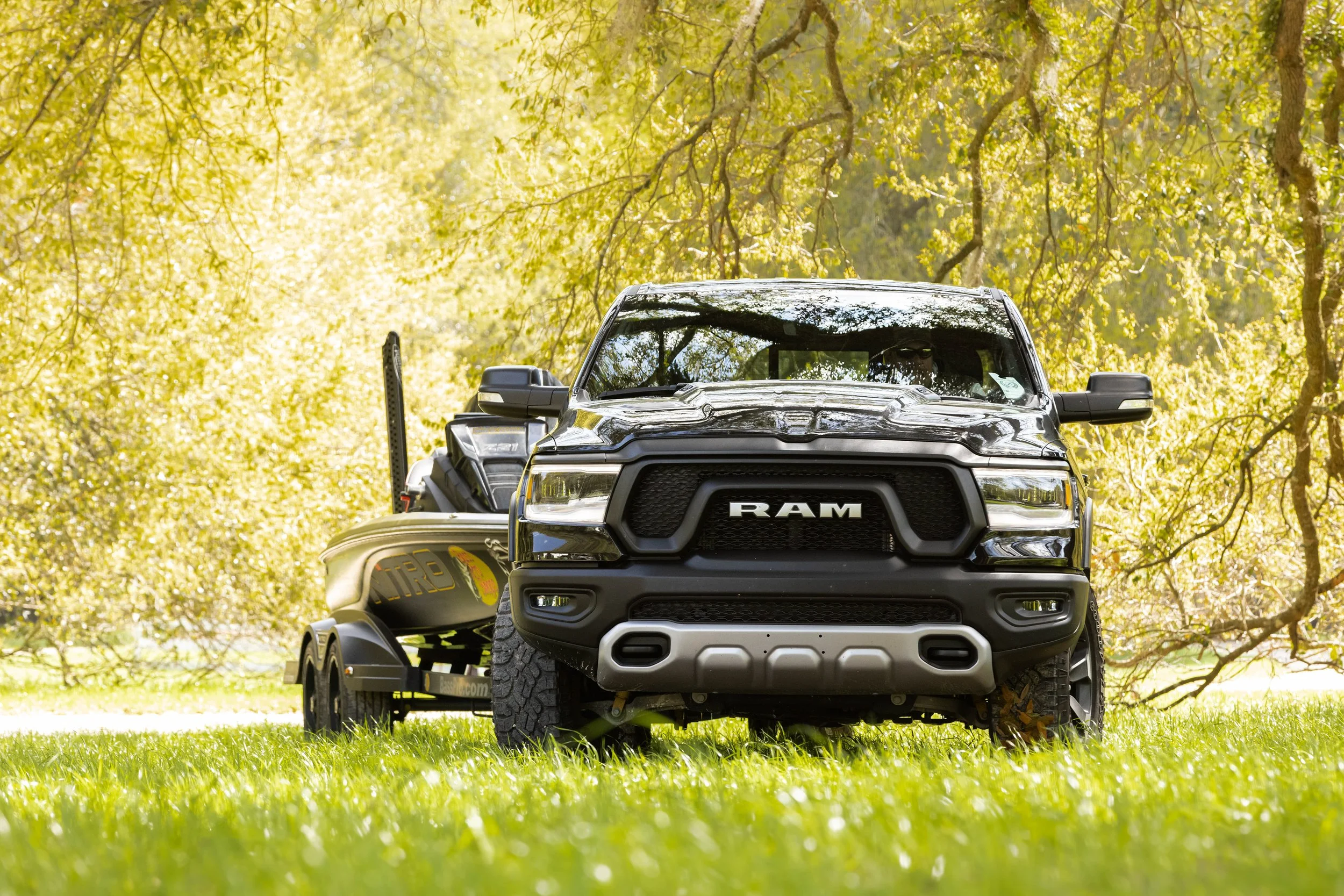 A black RAM pickup truck attached to a trailer with a jet boat, parked on a grassy field under a tree with green leaves.