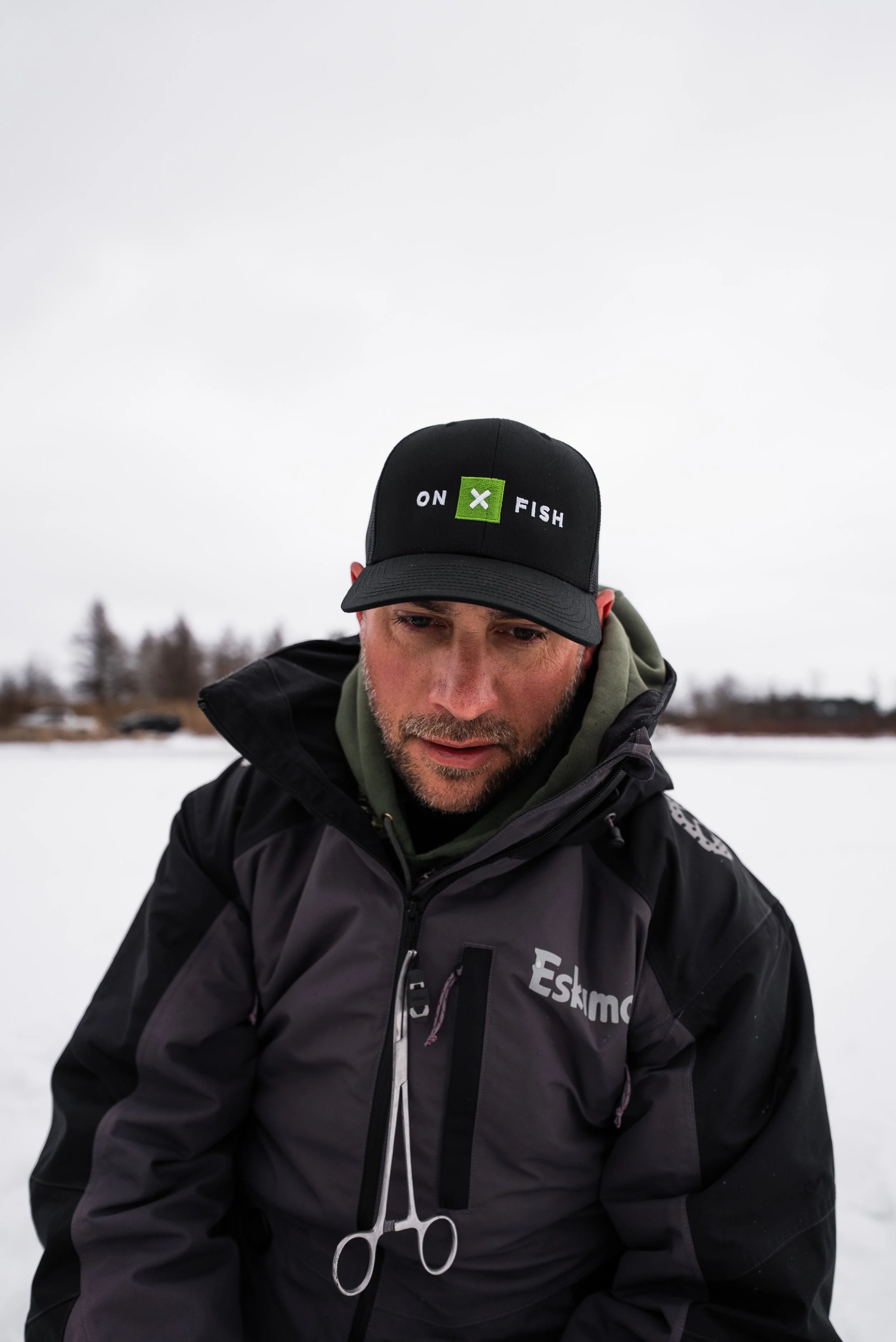 A man in a black jacket standing outdoors in a snowy landscape, wearing a black cap with 