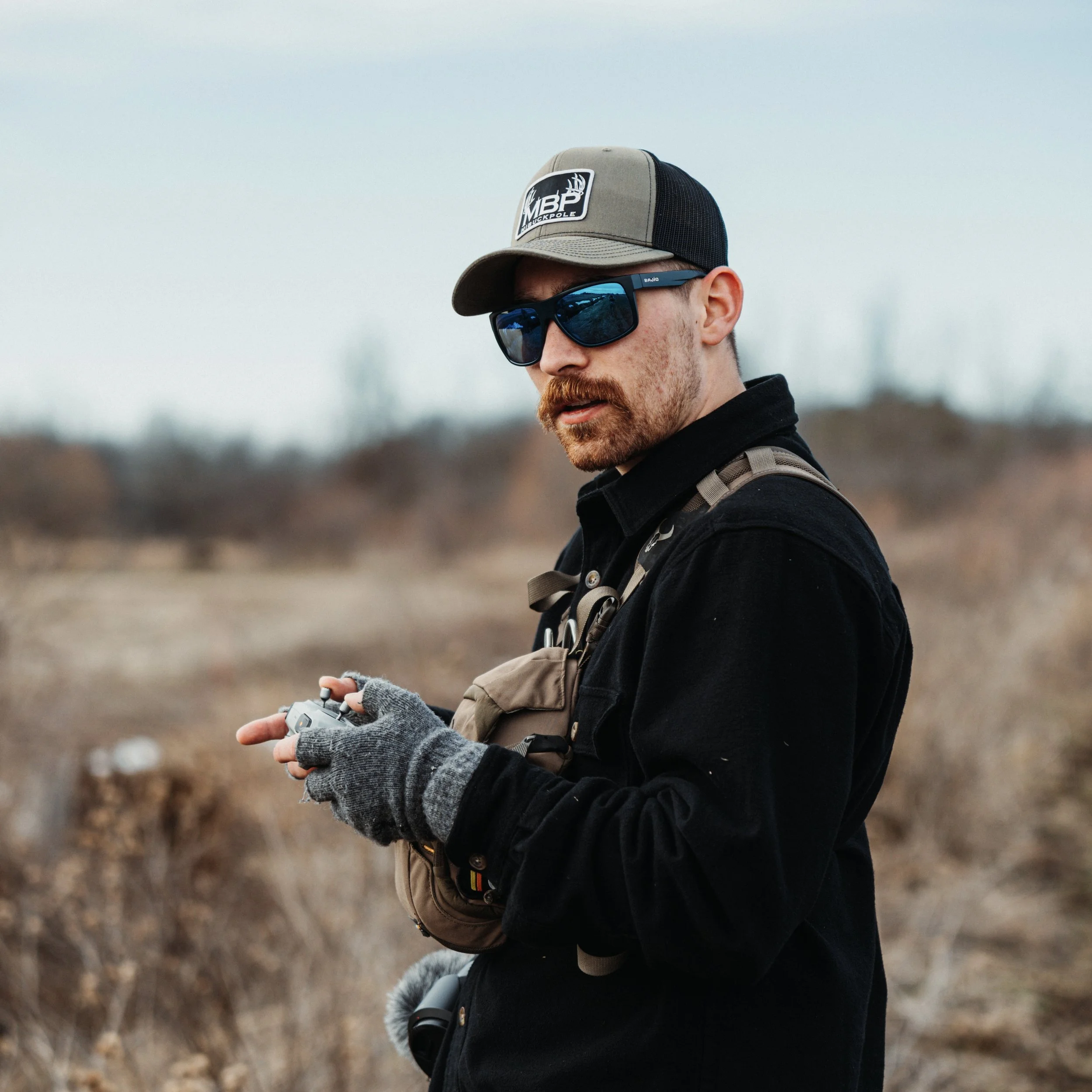 A man with a beard and sunglasses standing outdoors in a dry, bushy area, holding a remote control.