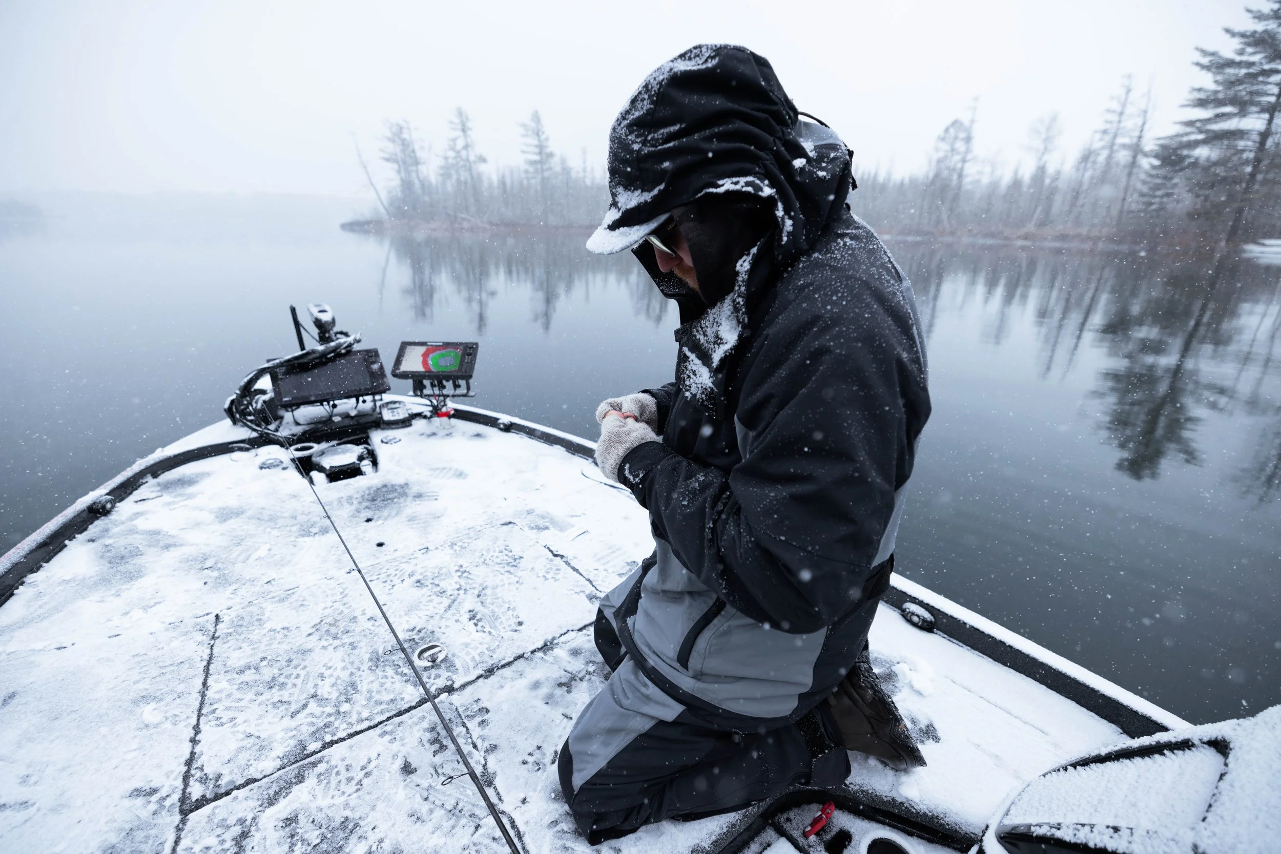 A person dressed warmly in winter gear is kneeling on a snow-covered boat at the edge of a frozen body of water, with snow falling around and a distant, foggy shoreline with trees.
