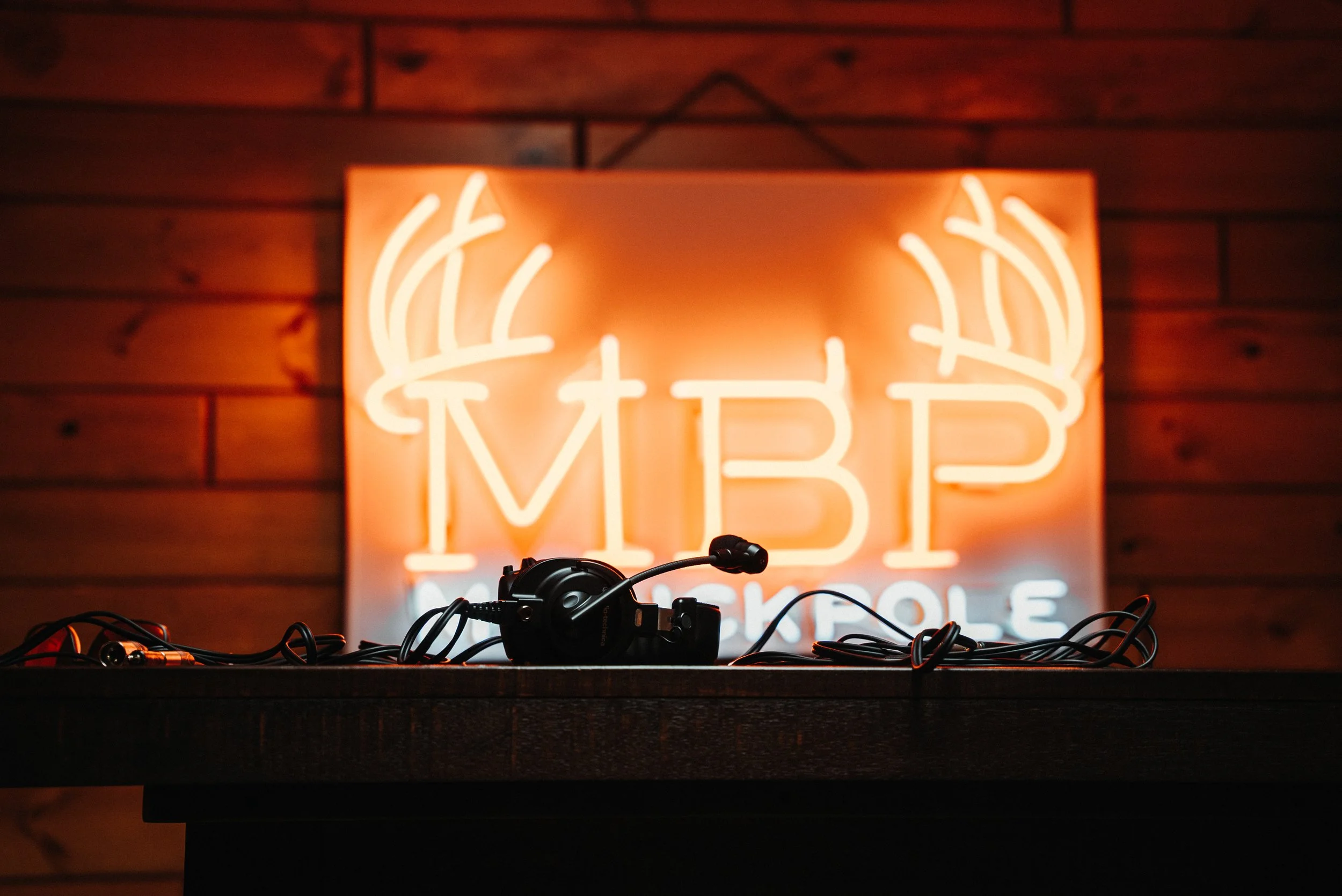 A neon sign with deer antlers and the letters 'MIB' on a wooden wall. In front, a DJ mixer and headphones are on a table.